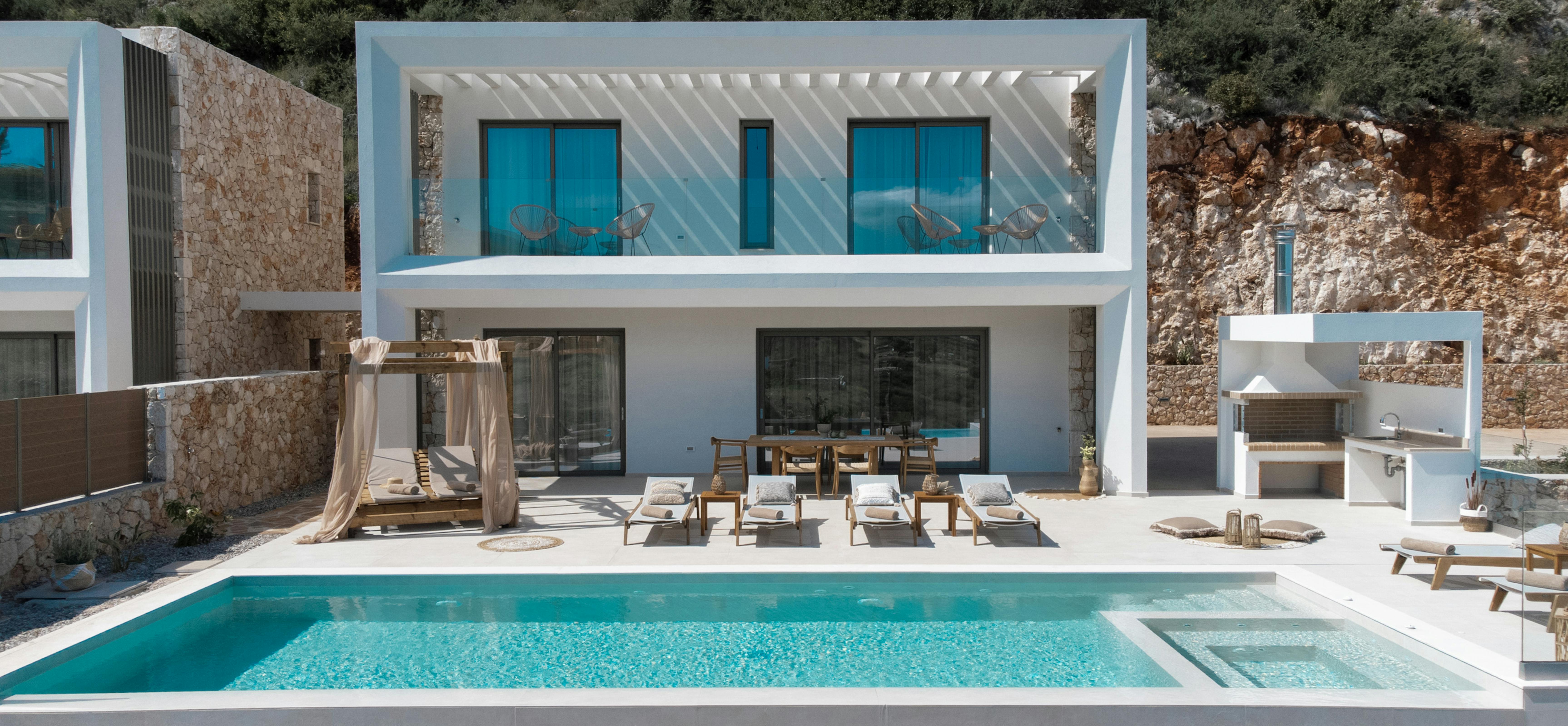 Modern white villa with two-story design, large windows, and infinity pool set against rocky hillside. Wooden outdoor furniture and outdoor kitchen complete the luxury retreat aesthetic.