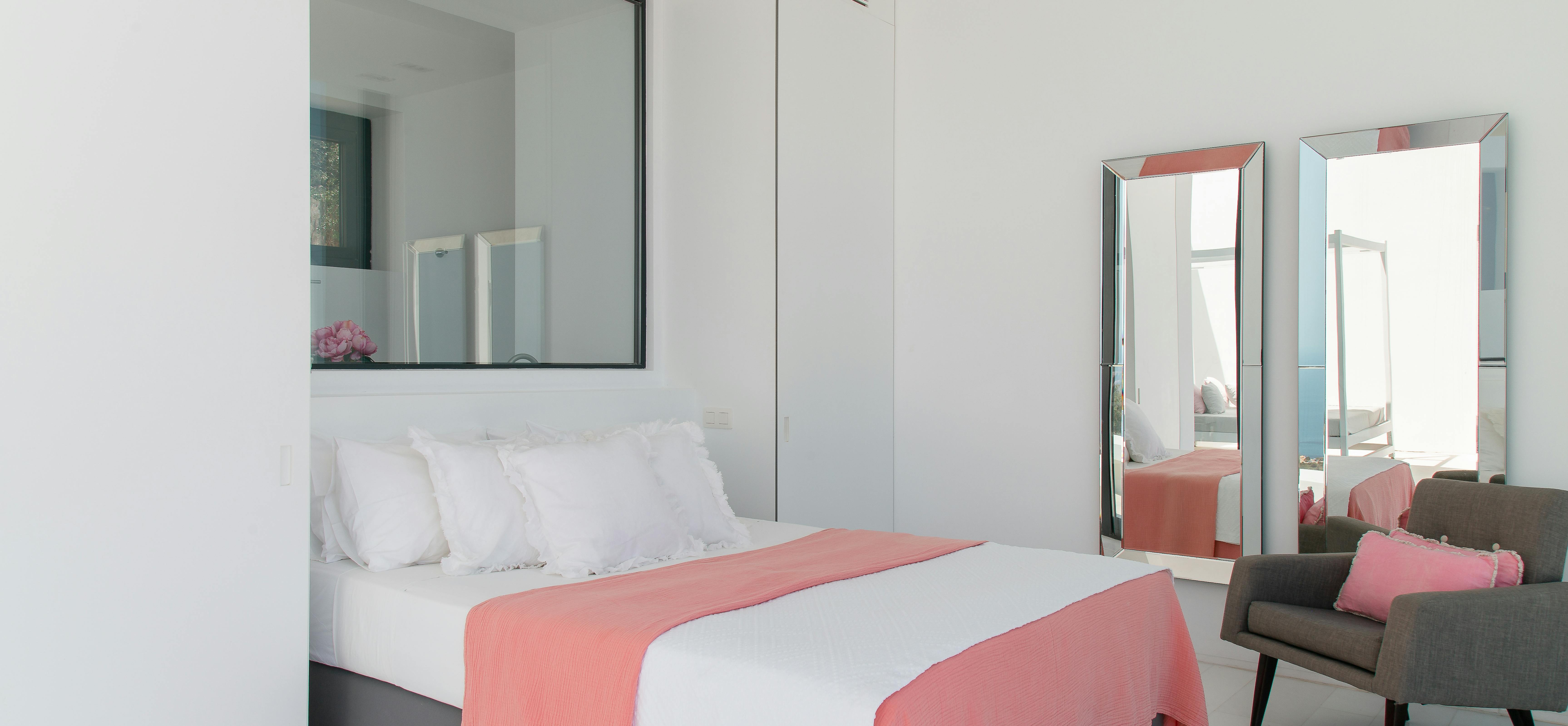 Modern bedroom with coral accent bedding, mirrored wardrobes, and grey upholstered chair overlooking terrace. Neutral palette with pops of pink creates serene atmosphere.