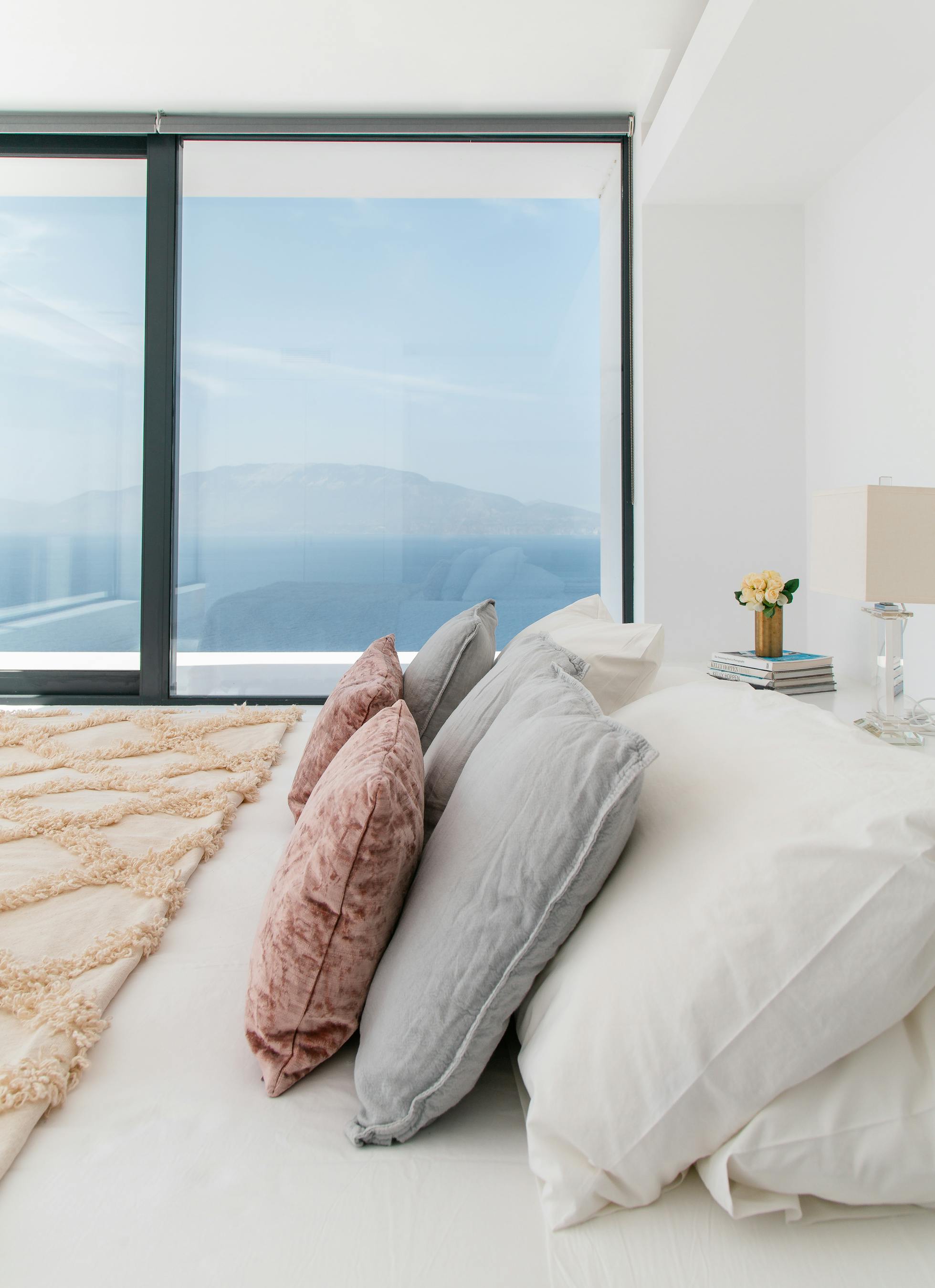 Luxurious bedroom with layered neutral bedding, pink accents, and expansive windows framing mountain and sea views. White interior emphasises natural beauty beyond