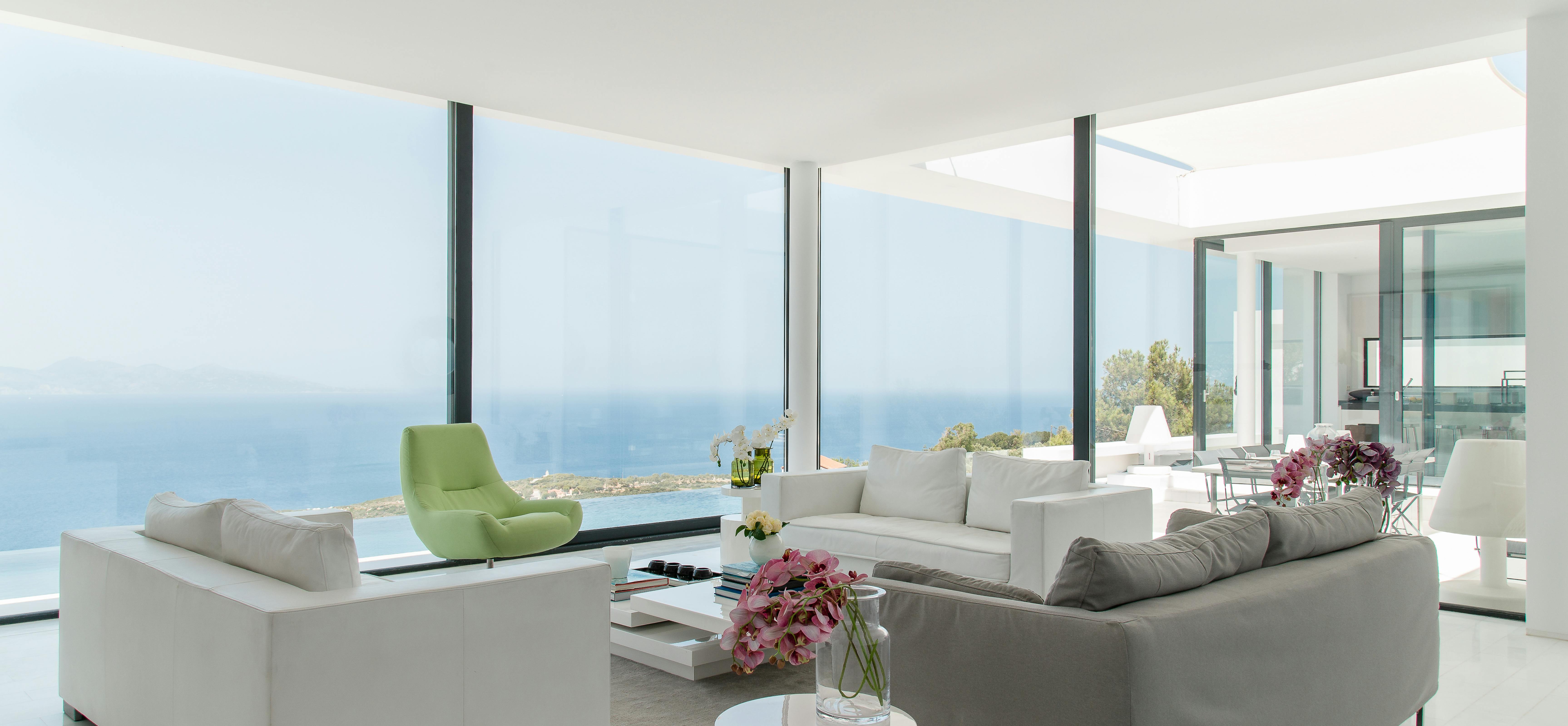 Open-plan living area with white and grey furniture, lime green accent chair, and wraparound windows showcasing endless ocean views. Pink flowers soften the modern aesthetic.
