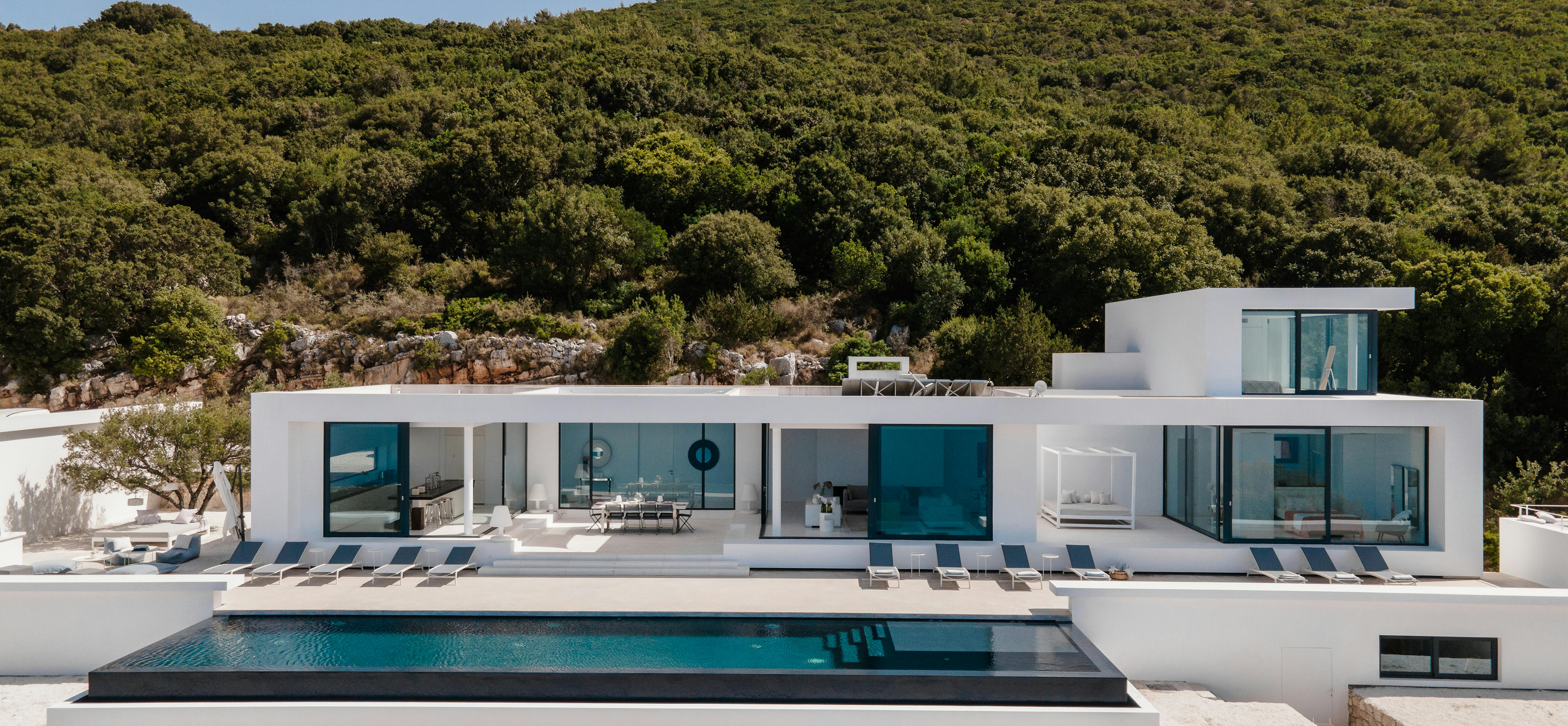 Modern white villa with geometric architecture featuring floor-to-ceiling glass walls and black-framed infinity pool set against forested hillside. Clean minimalist design emphasises indoor-outdoor living.