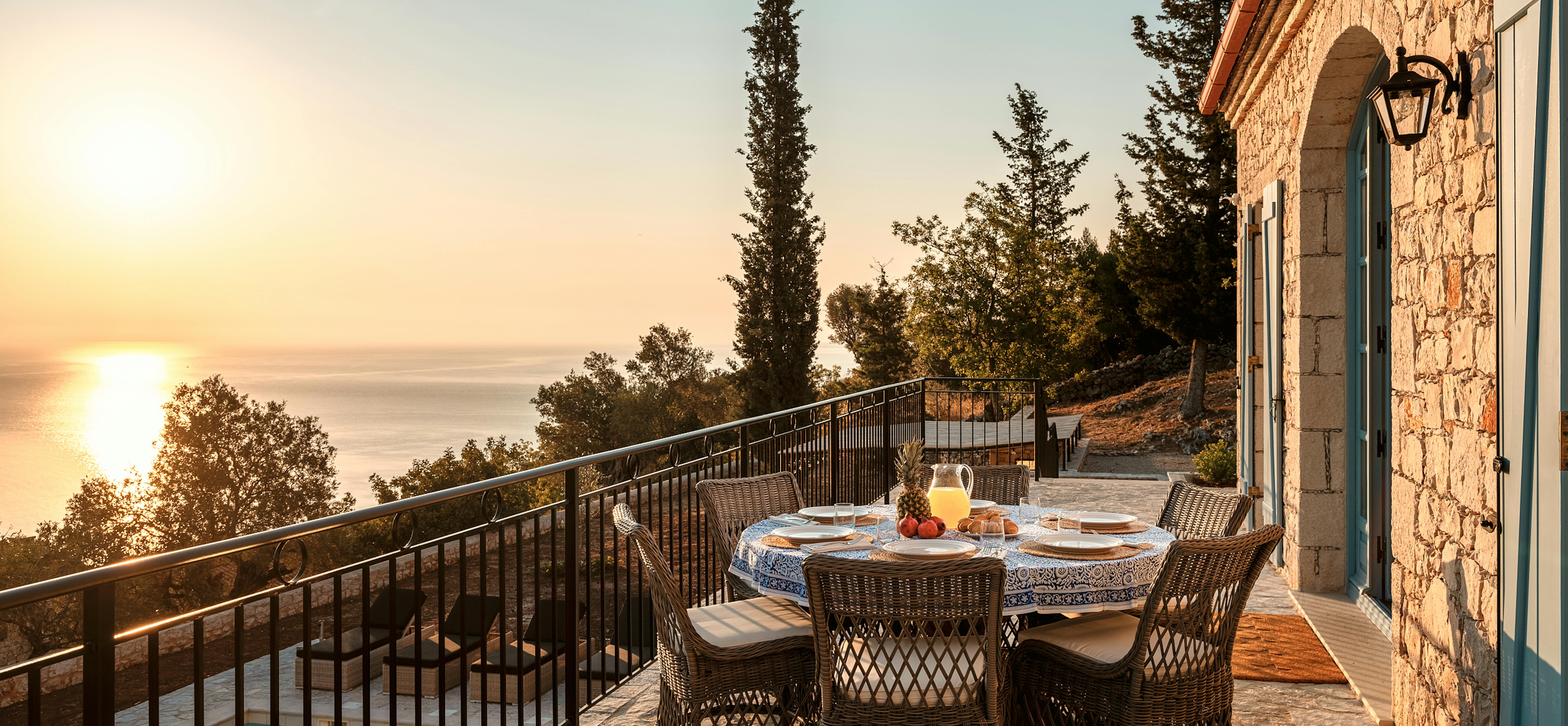 Stone terrace dining area at golden hour with woven chairs around table set for breakfast overlooking infinity pool and sea. Traditional Mediterranean architecture with turquoise shutters frames the scenic view.