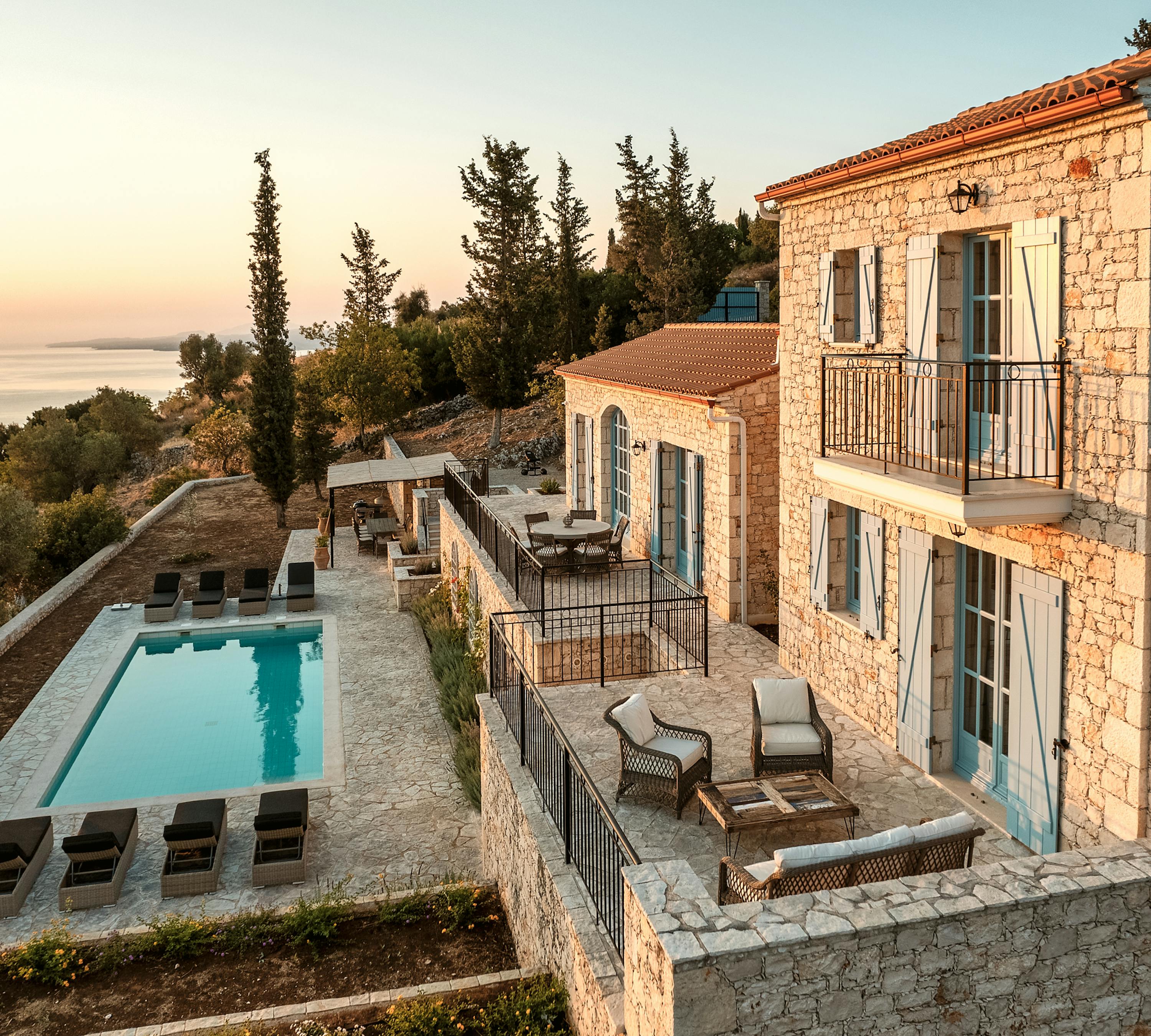 Traditional Mediterranean stone villa with terracotta roof overlooking sea at sunset, featuring multiple terraces, infinity pool, and turquoise shutters. Warm golden light illuminates the limestone architecture and cypress trees.