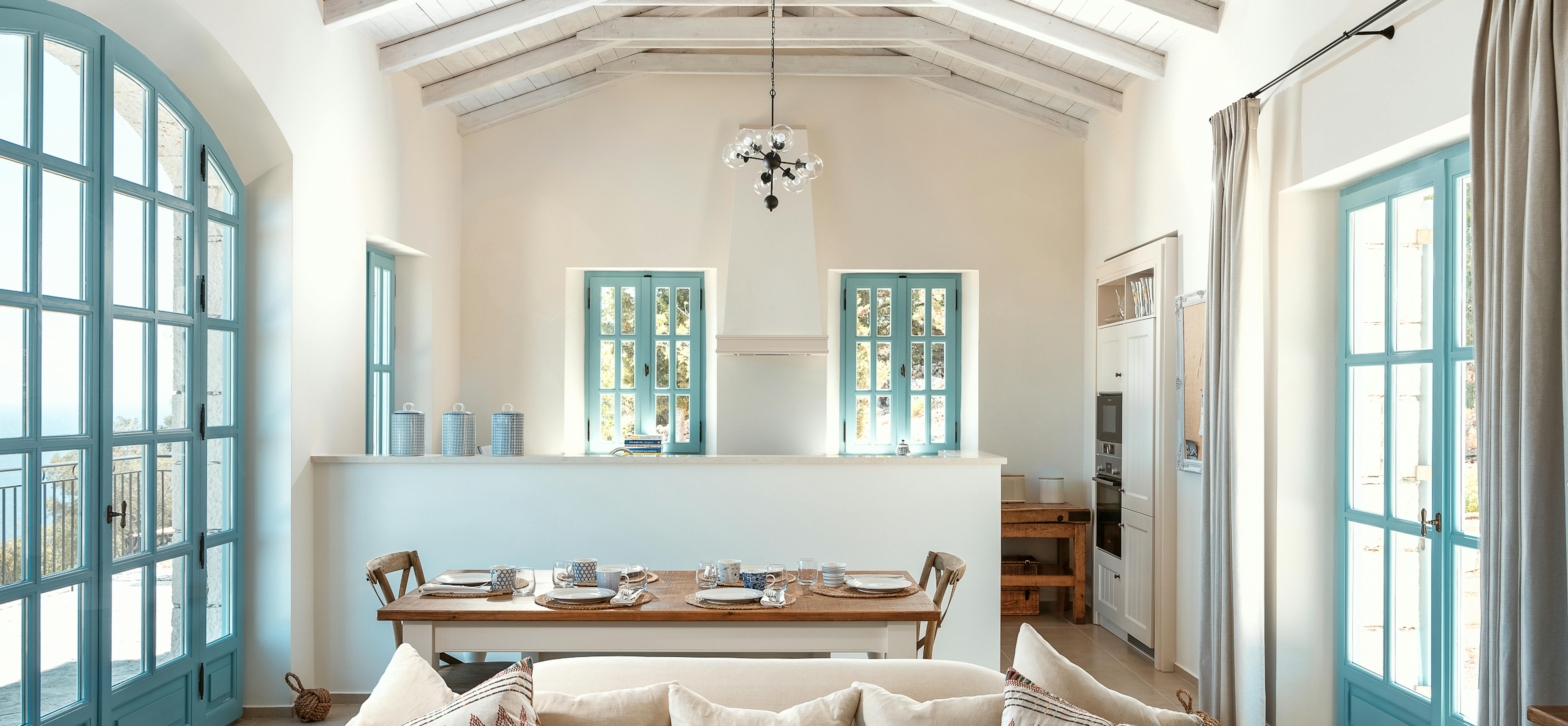 Open-plan living space with cathedral ceiling, turquoise arched windows, and dining area overlooking kitchen. Exposed beams, neutral sofa with geometric pillows, and traditional Mediterranean details define the interior.