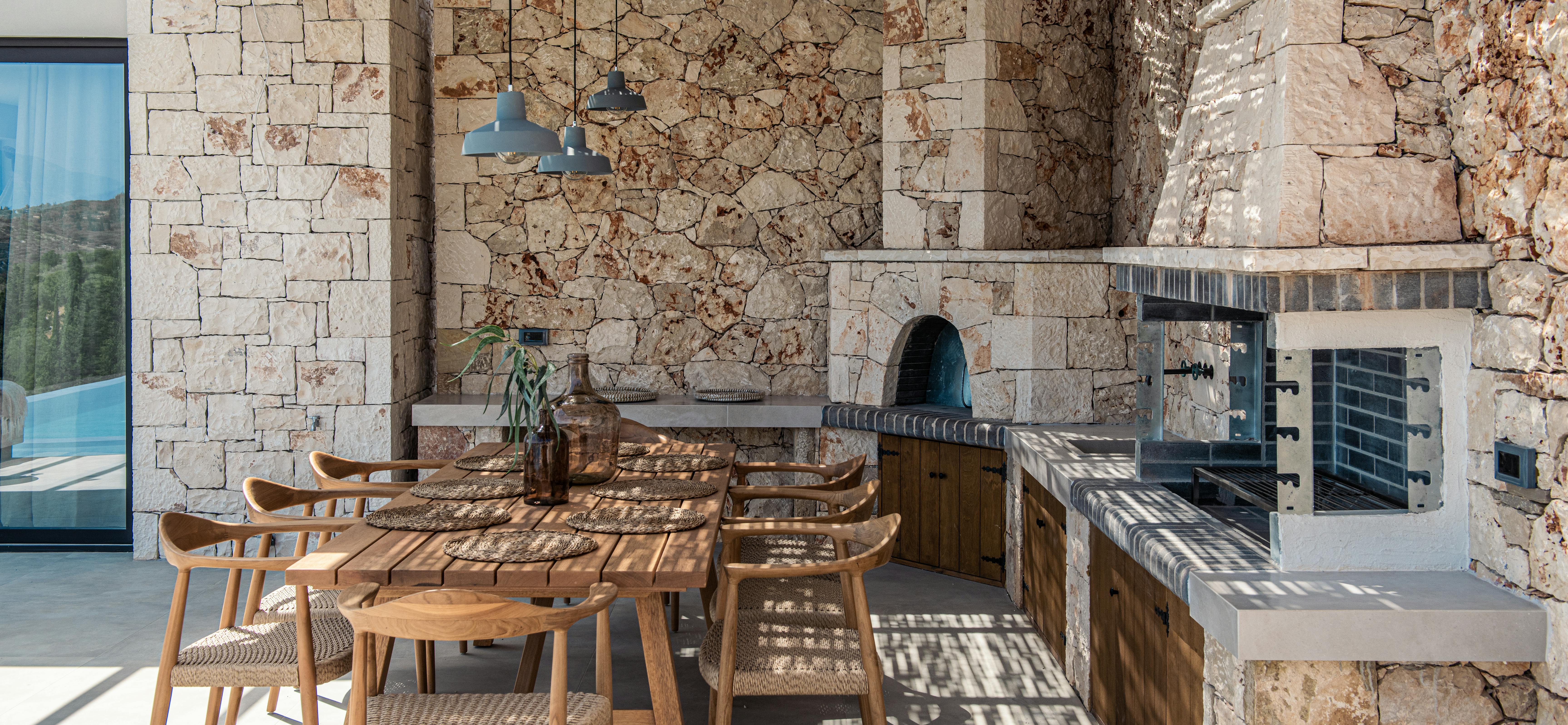 Stone-walled outdoor kitchen with wood-fired oven, wooden dining table, and rustic Mediterranean design.