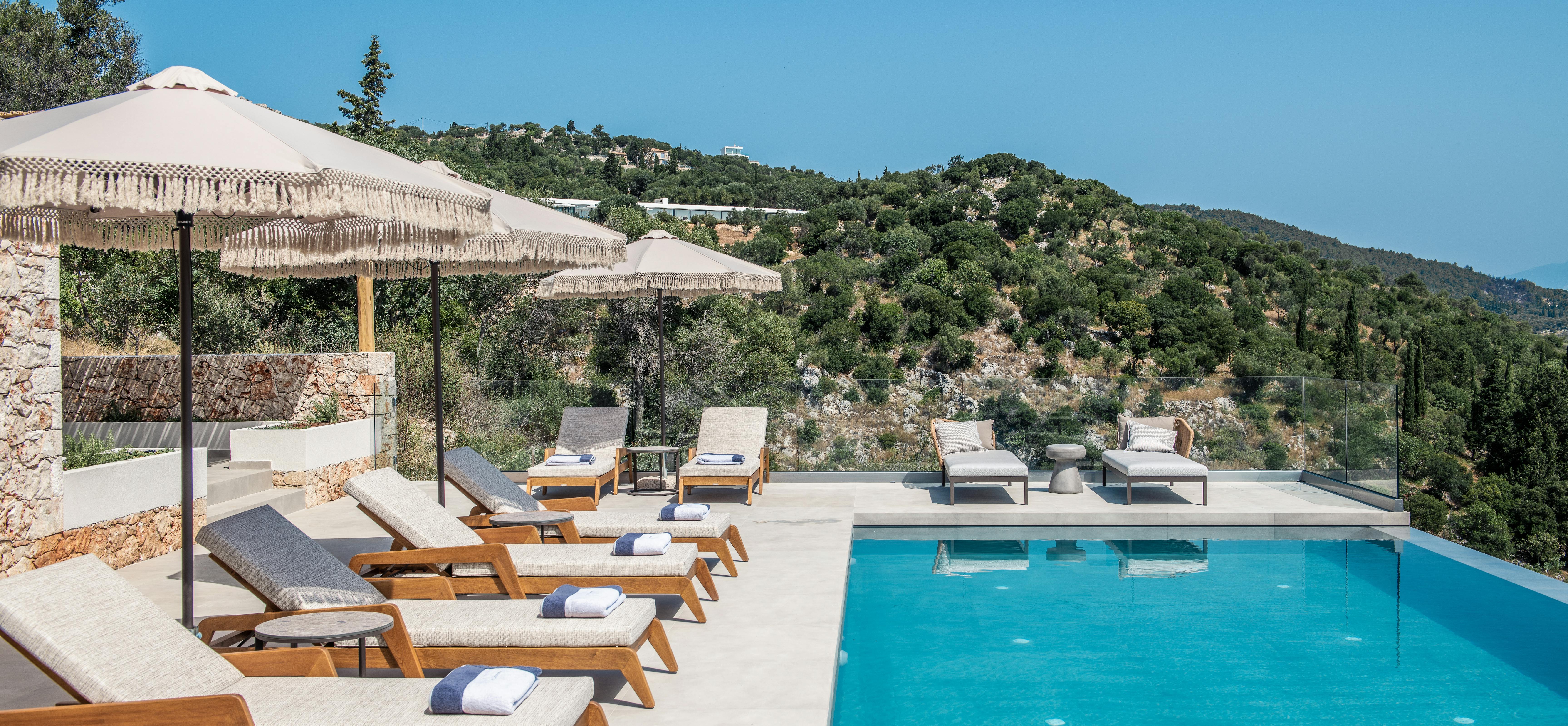 Luxury pool terrace with wooden loungers, fringe umbrellas, and panoramic Mediterranean hillside views.
