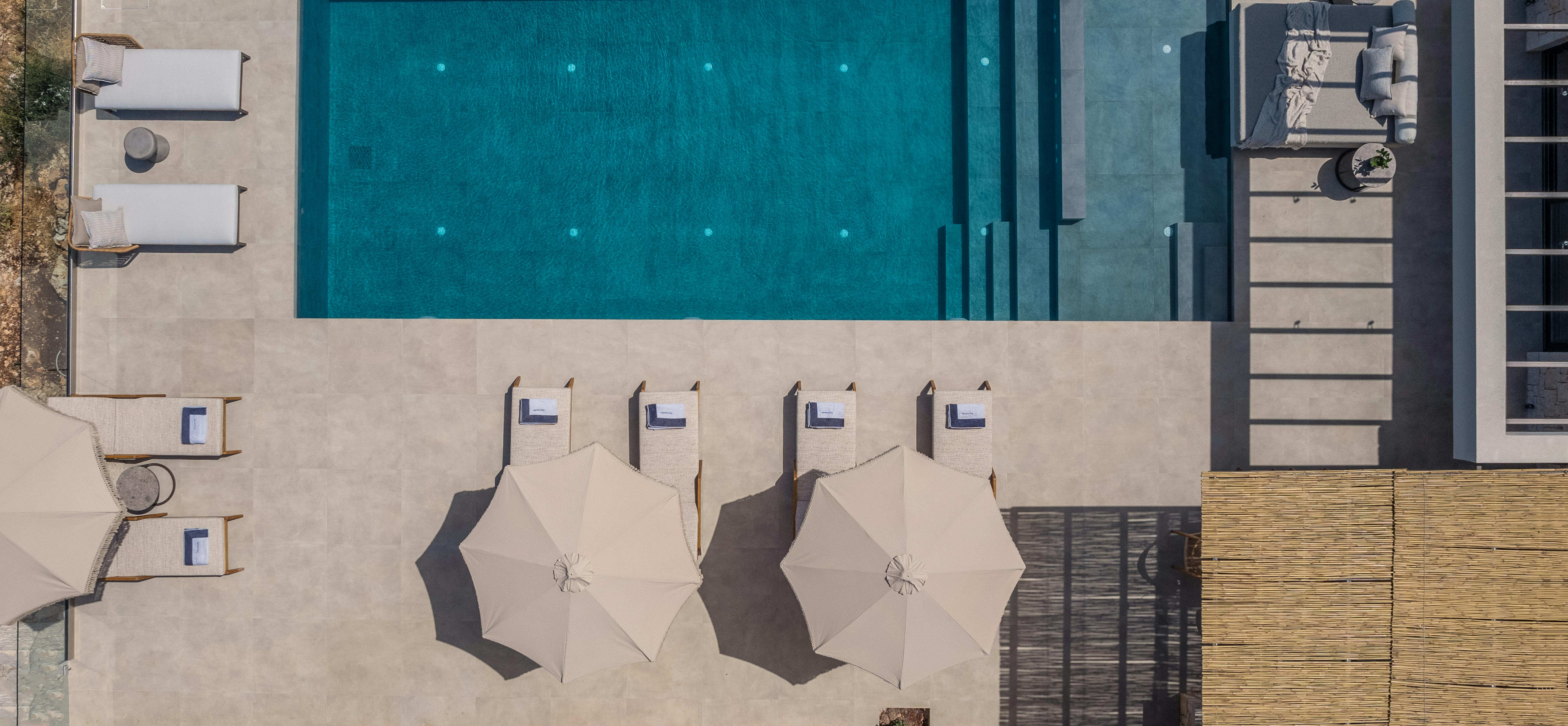 Aerial view of modern pool with glass barriers, beige umbrellas, and geometric deck loungers.