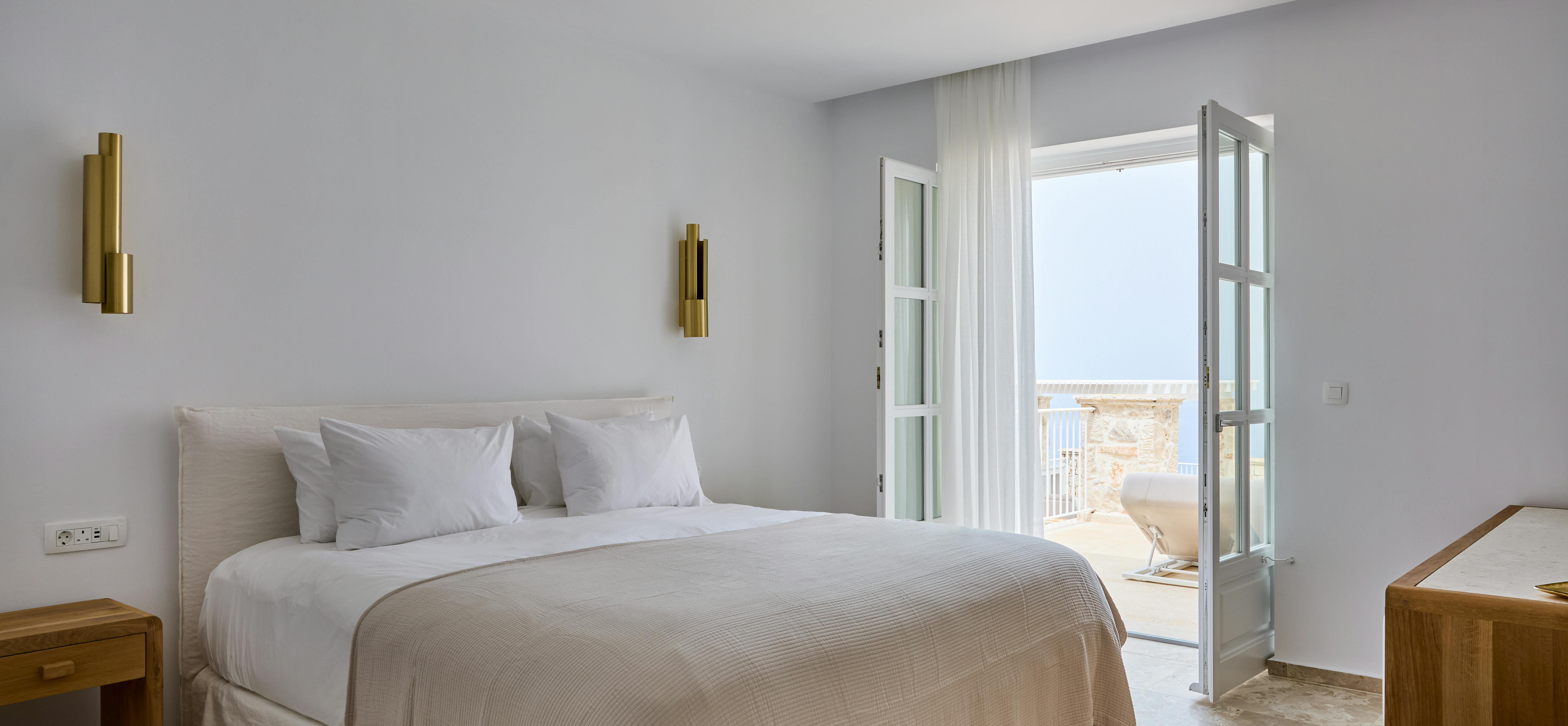 A master bedroom maintains the villa's neutral palette with white linens and brass wall sconces. French doors open to a private balcony overlooking the Mediterranean coastline.