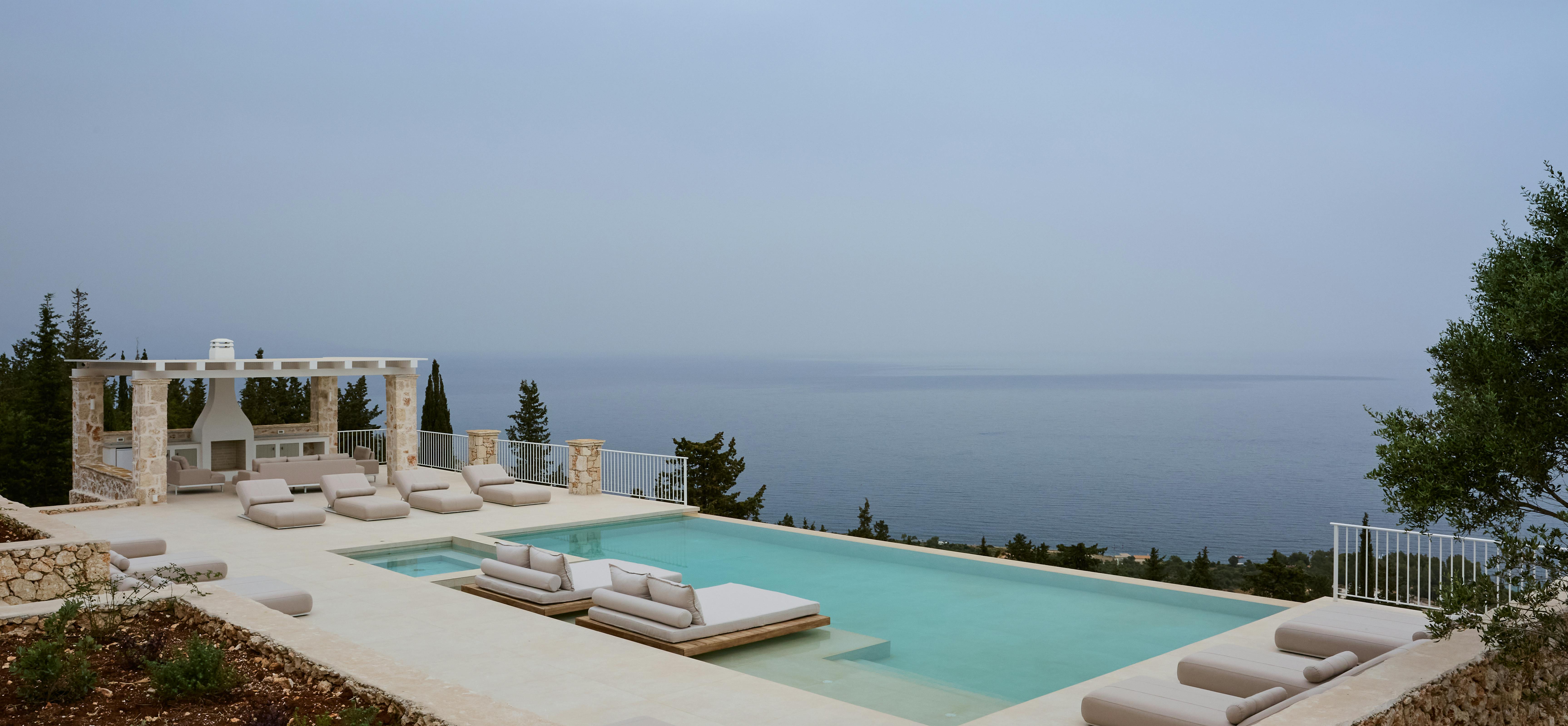 A spectacular pool overlooking the Mediterranean Sea features plush neutral loungers and a separate spa pool. The clifftop terrace includes a stone pergola structure with panoramic ocean views stretching to the horizon.