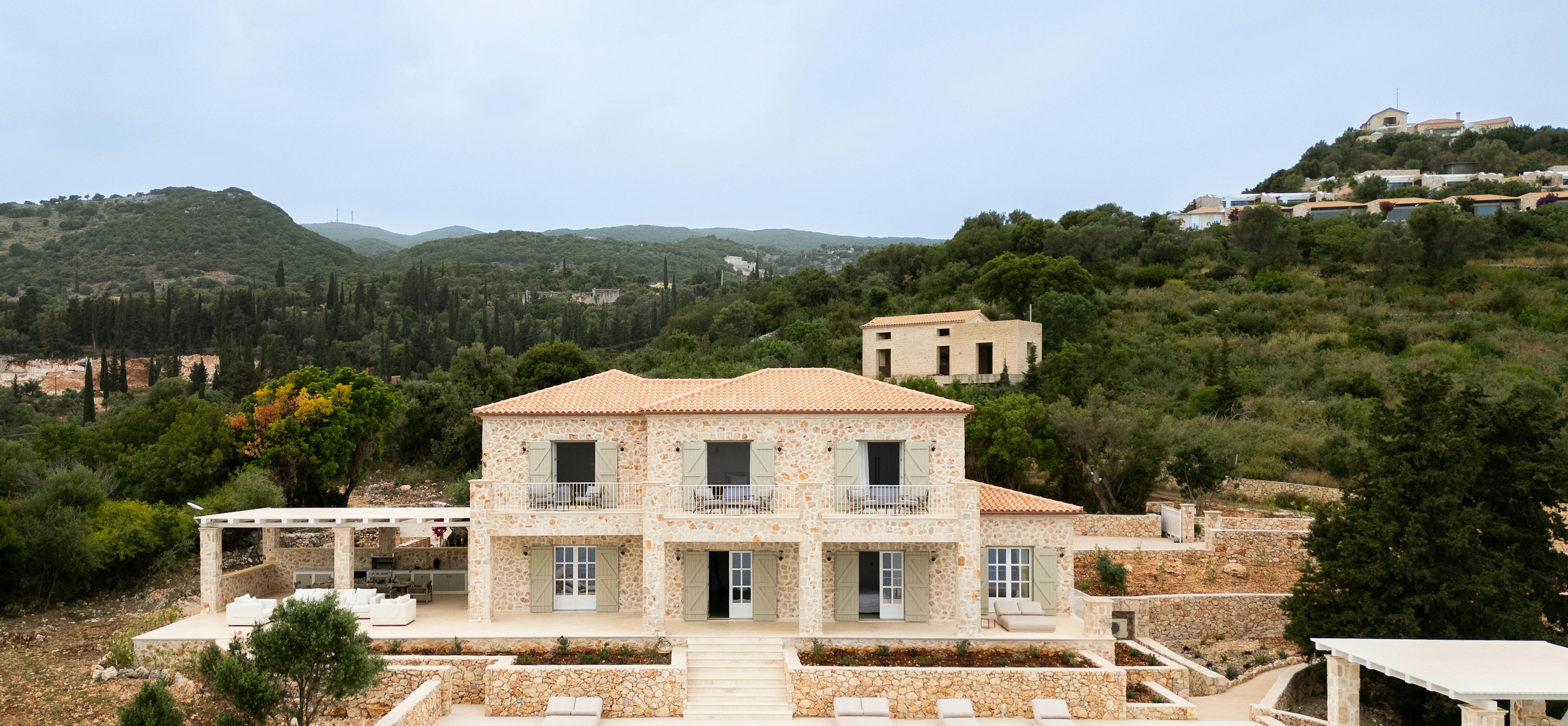 A Mediterranean-style stone villa with terracotta roof tiles sits among rolling hills, featuring a pristine swimming pool with white loungers and terraced stone walls. The property blends traditional architecture with modern luxury amenities in a scenic hillside setting.