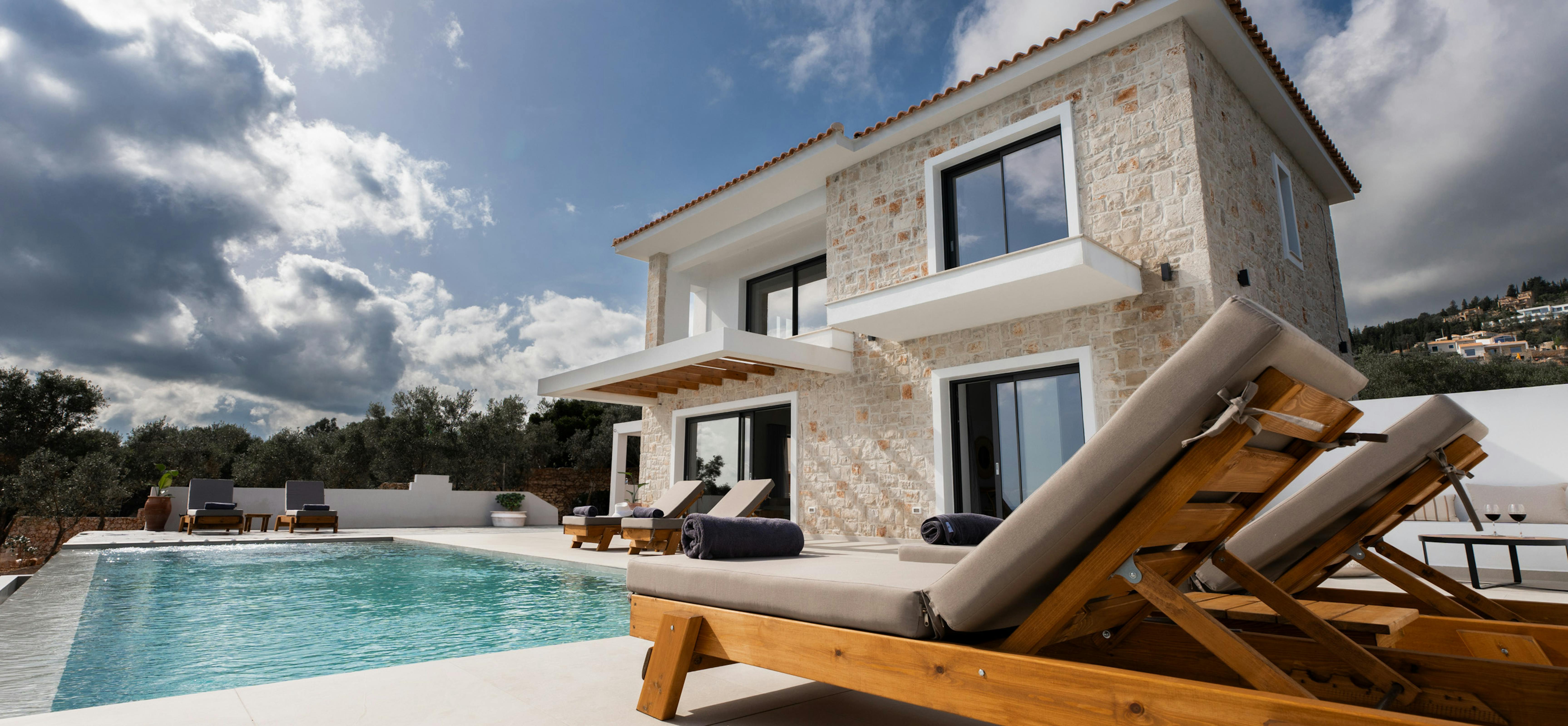 A modern Mediterranean villa with natural stone walls overlooks a swimming pool with timber sun loungers arranged on the deck. The two-storey home features contemporary design elements against a dramatic cloudy sky.