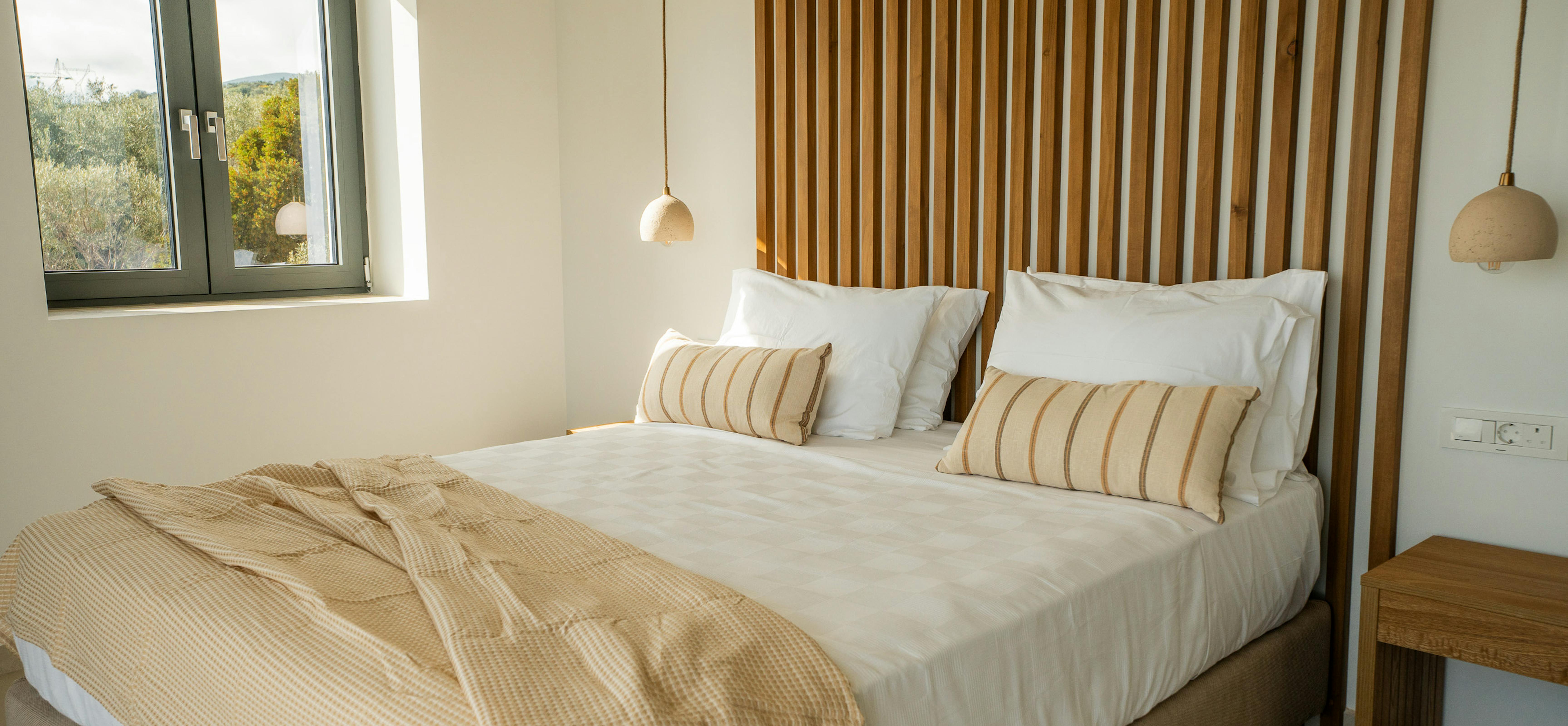 The bedroom's timber slat headboard creates a contemporary focal point with integrated pendant lighting. White linens and striped cushions maintain the neutral palette whilst large windows provide natural illumination.