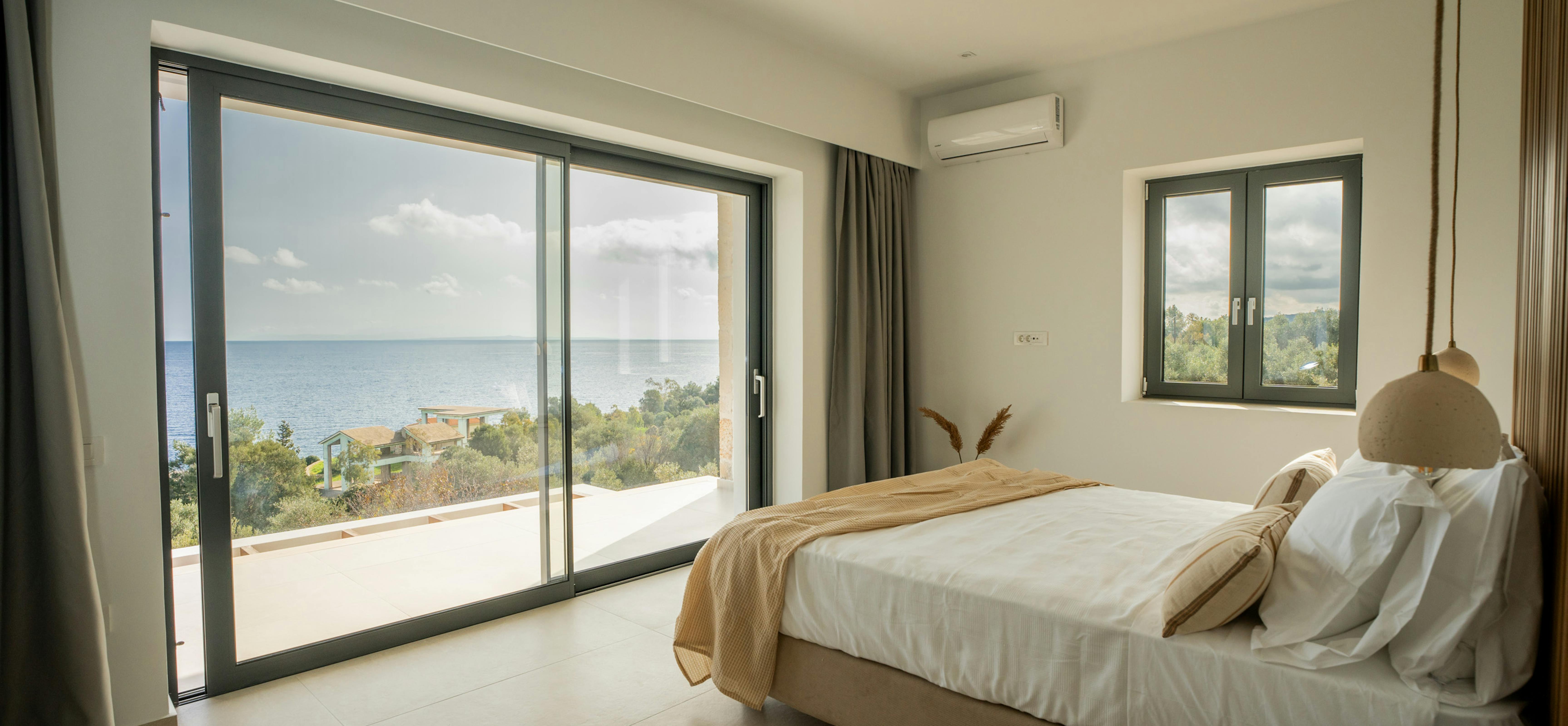 A minimalist bedroom with ocean views features large sliding doors opening to a private terrace. The neutral palette includes white linens, timber furniture, and a woven pendant light with Mediterranean coastline visible beyond.