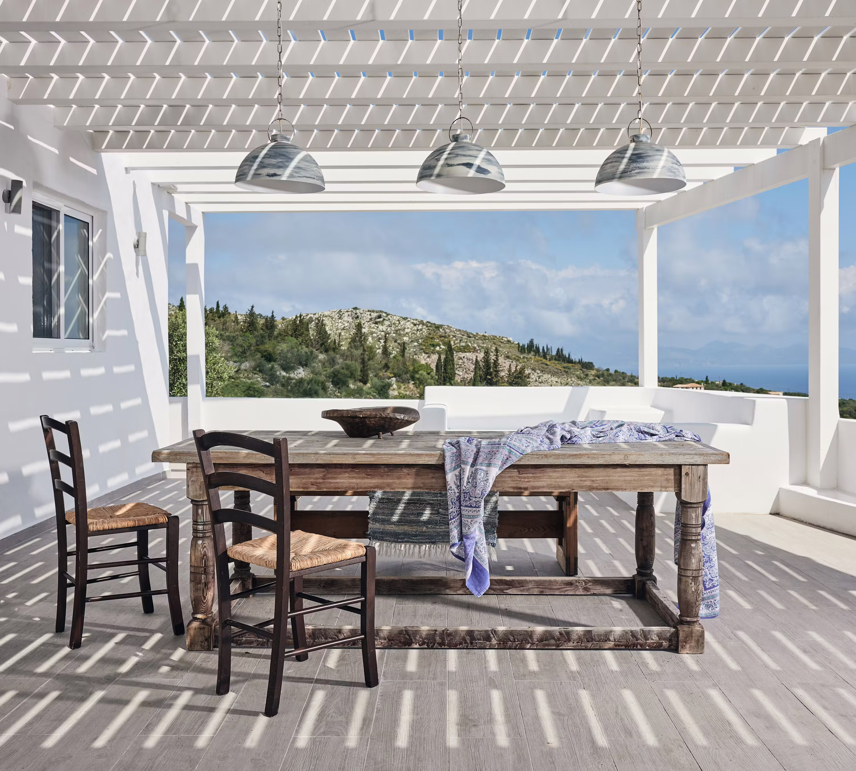 Traditional rush-seated chairs surround a rustic timber dining table on a covered terrace with industrial pendant lighting. The white pergola structure frames views of the Mediterranean landscape and creates dramatic shadow patterns across the deck.