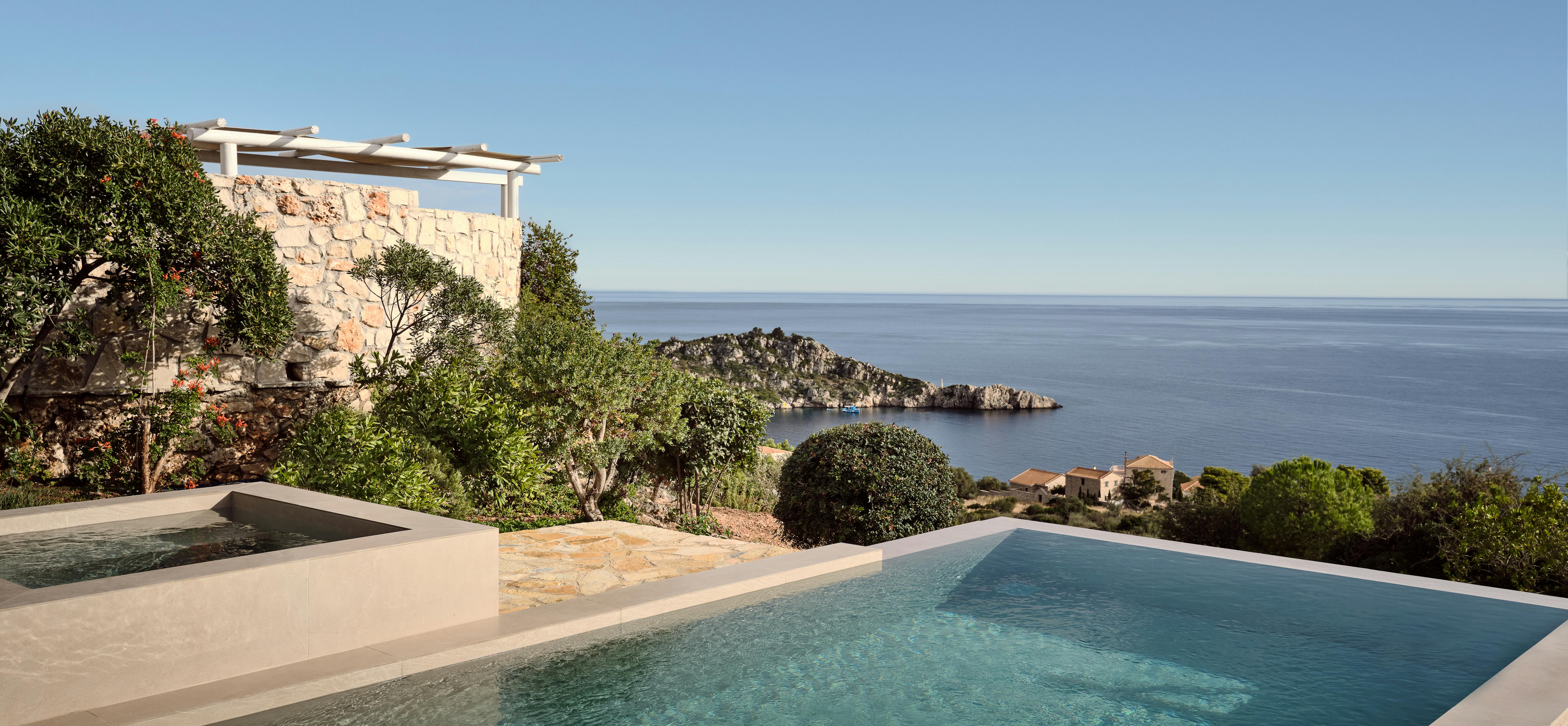 An infinity pool appears to merge with the Mediterranean Sea beyond, set within terraced stone walls and lush vegetation. The modern pool design contrasts beautifully with the traditional stone architecture and natural coastal landscape.