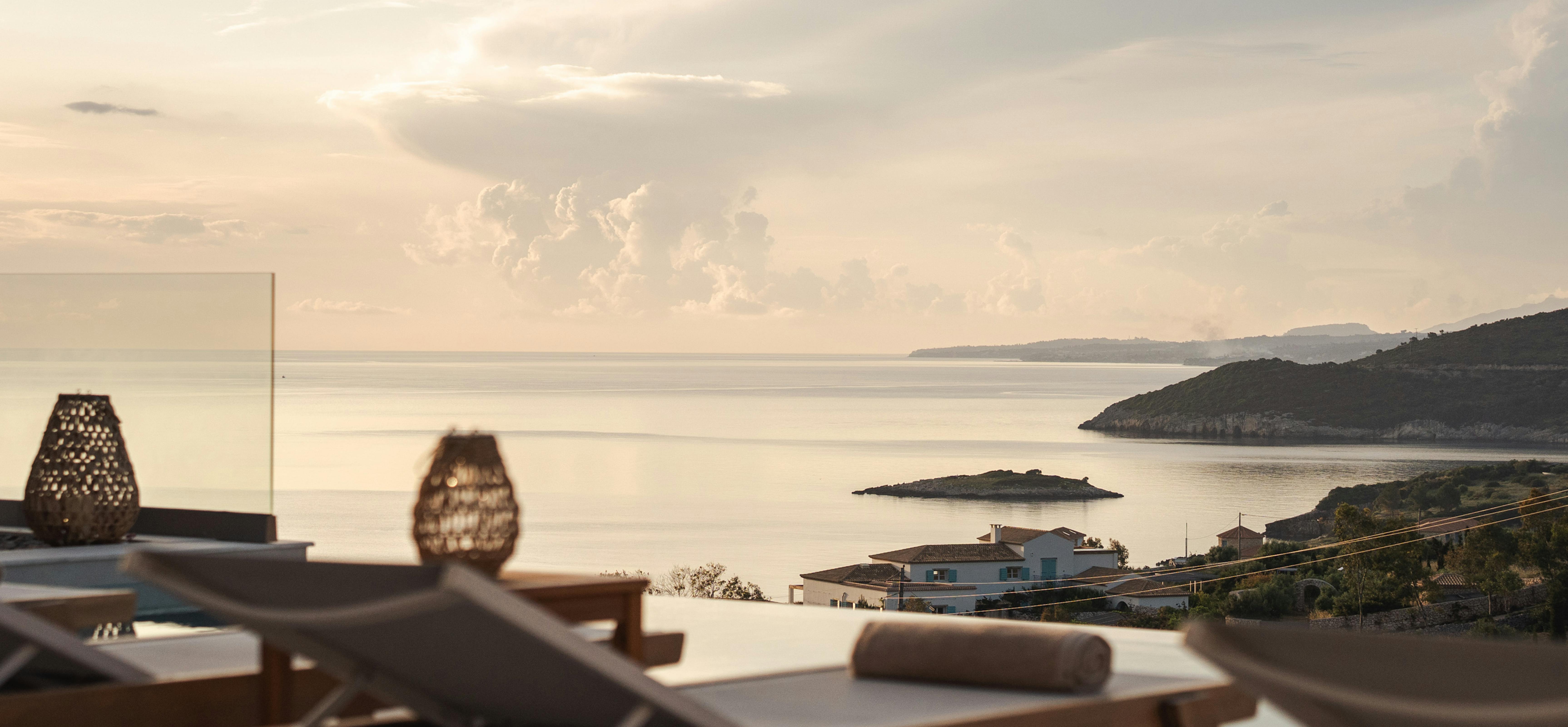 Wooden lounge chairs on terrace overlooking calm sea with dramatic cloudy sky at sunset.