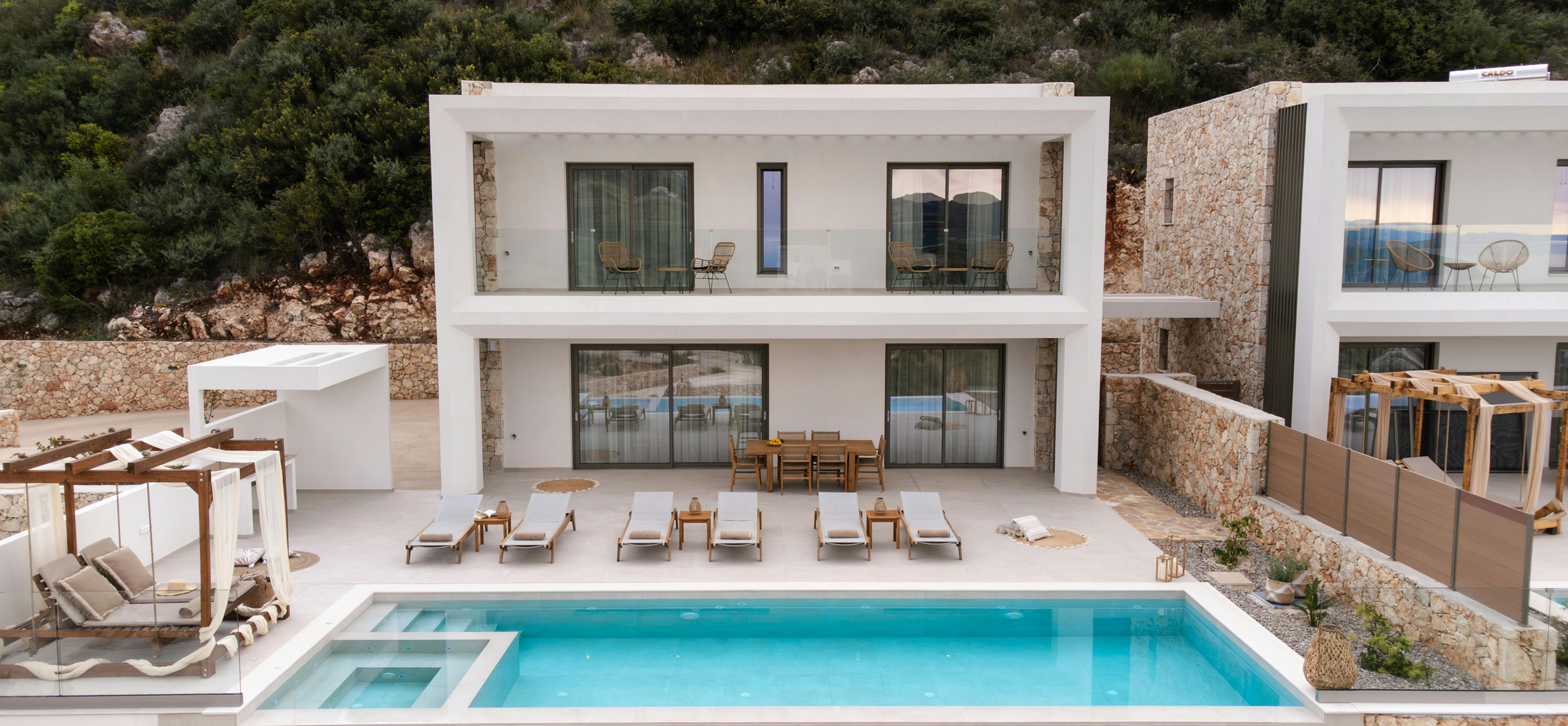Two-story white villa with pool, stone retaining walls, and forested hillside setting.