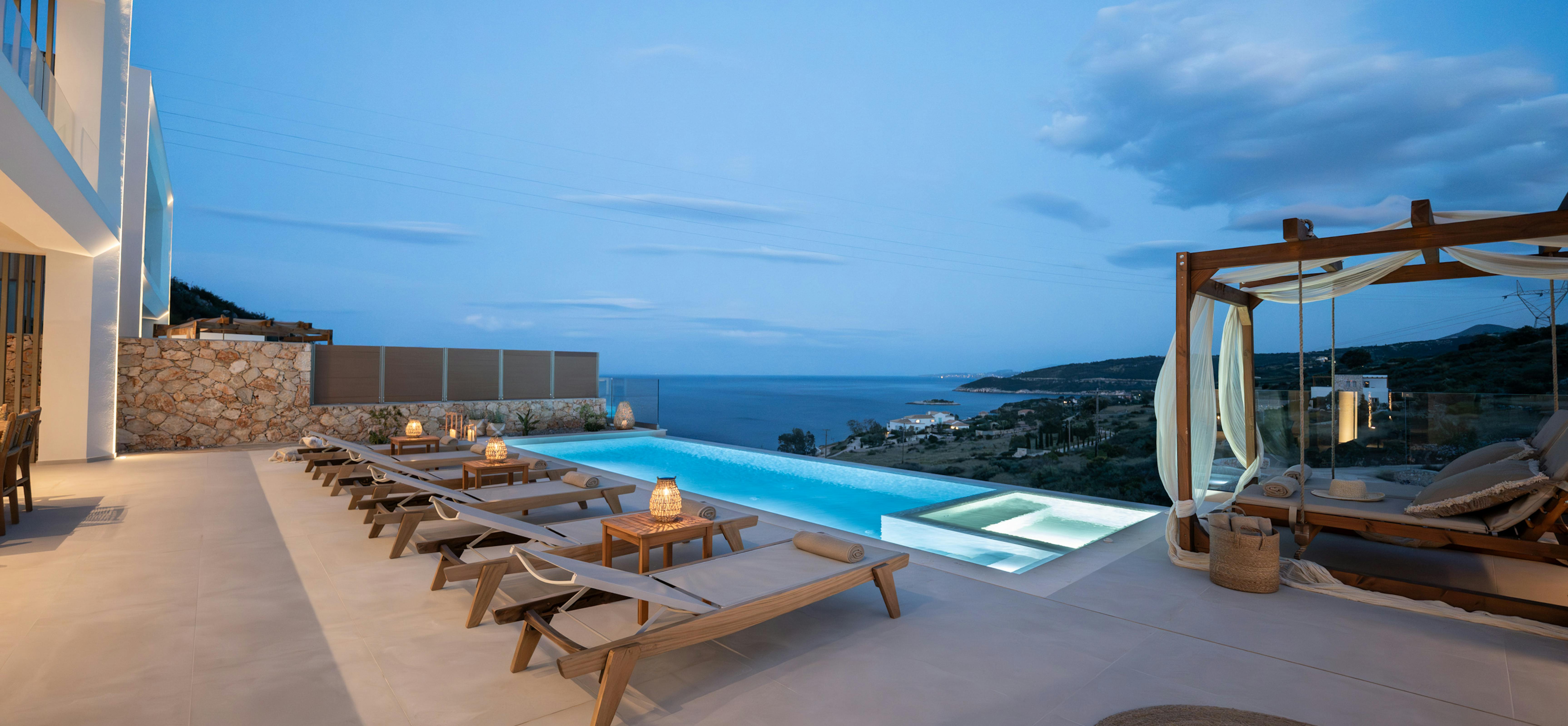 Evening pool terrace with illuminated infinity pool and panoramic Mediterranean coastline views.
