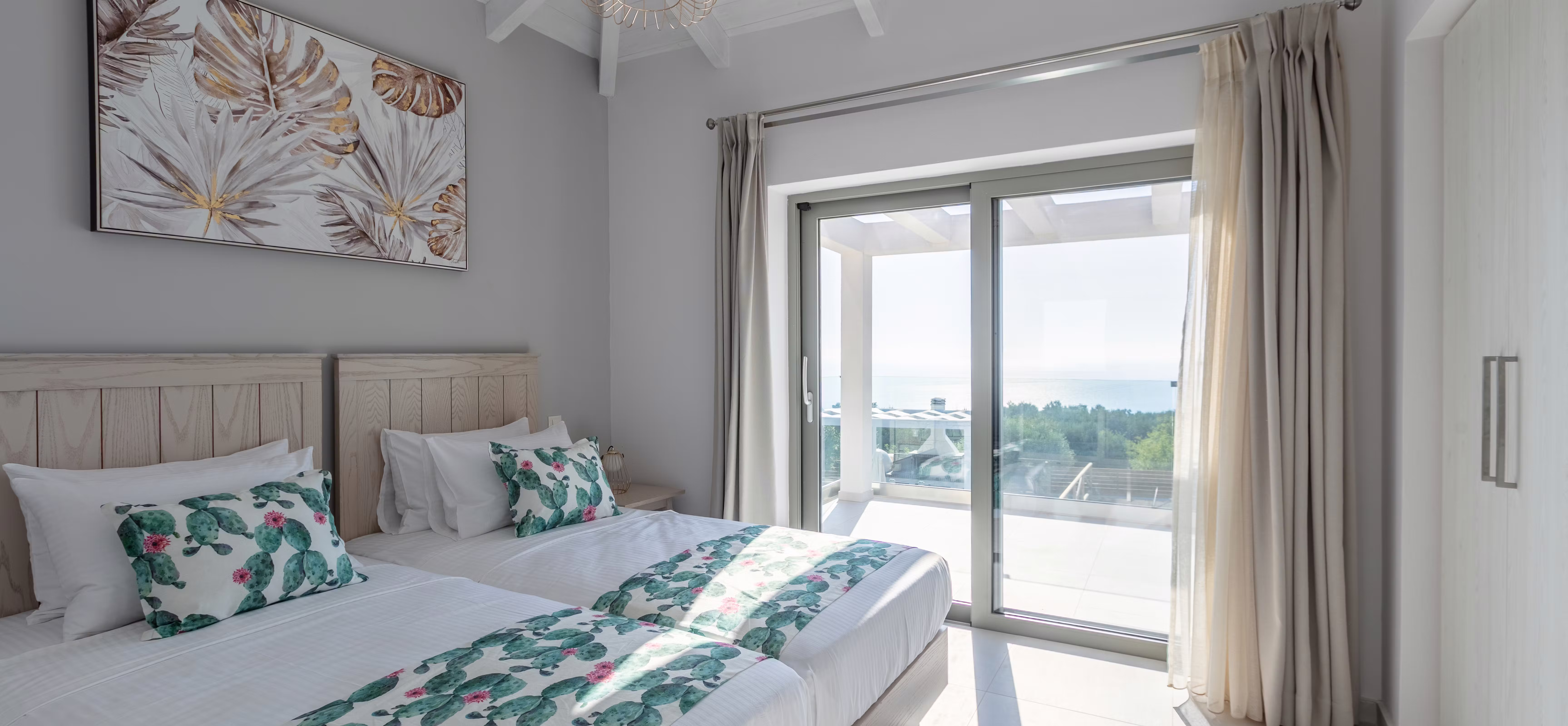 Bedroom with vaulted ceiling, wicker pendant light, tropical leaf artwork, bed with green floral bedding, white furnishings, and sliding doors opening to sunny terrace.