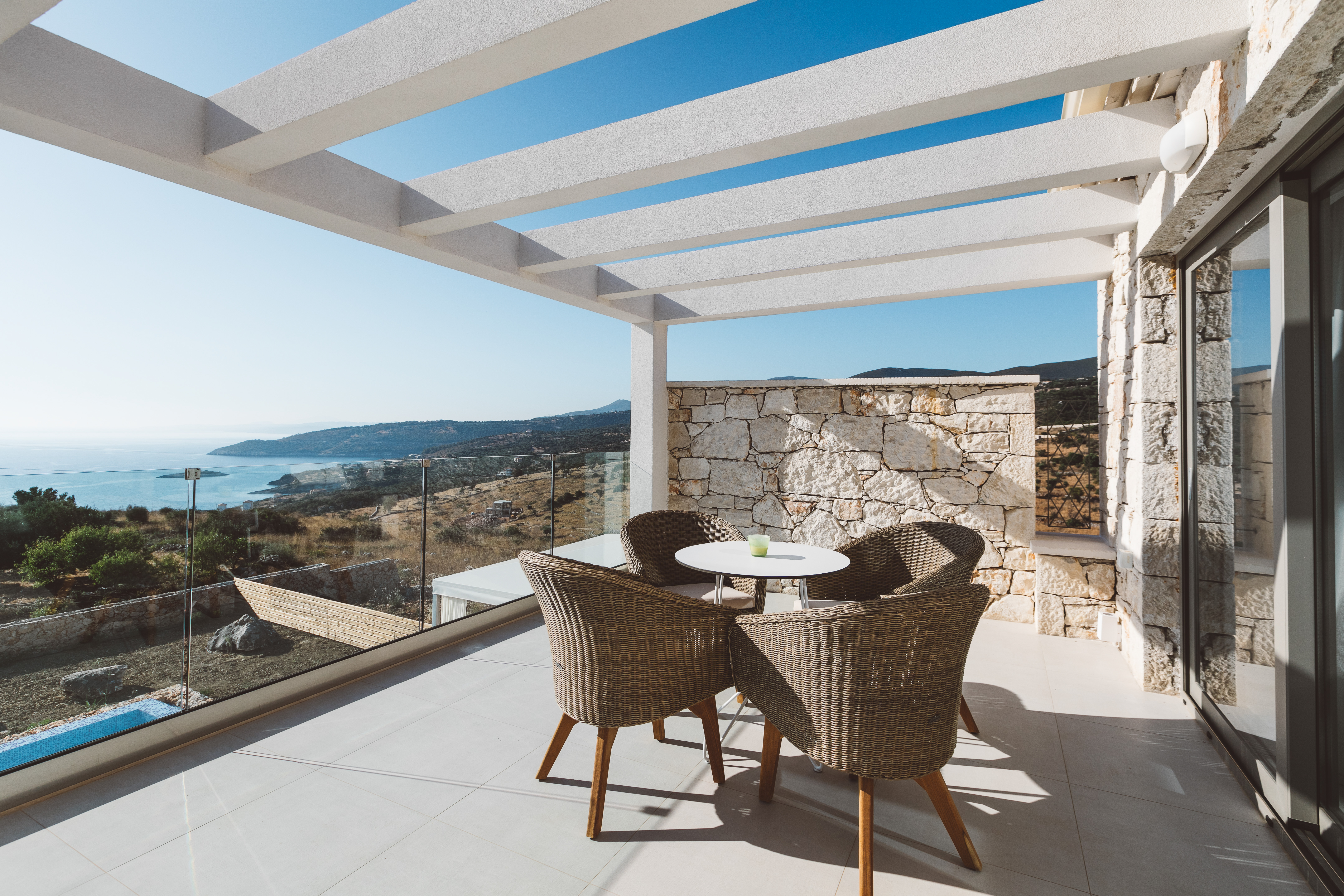 Covered terrace dining area with white pergola casting geometric shadows, wicker chairs around white table, stone accent wall, and glass-railed views of coastal hills.