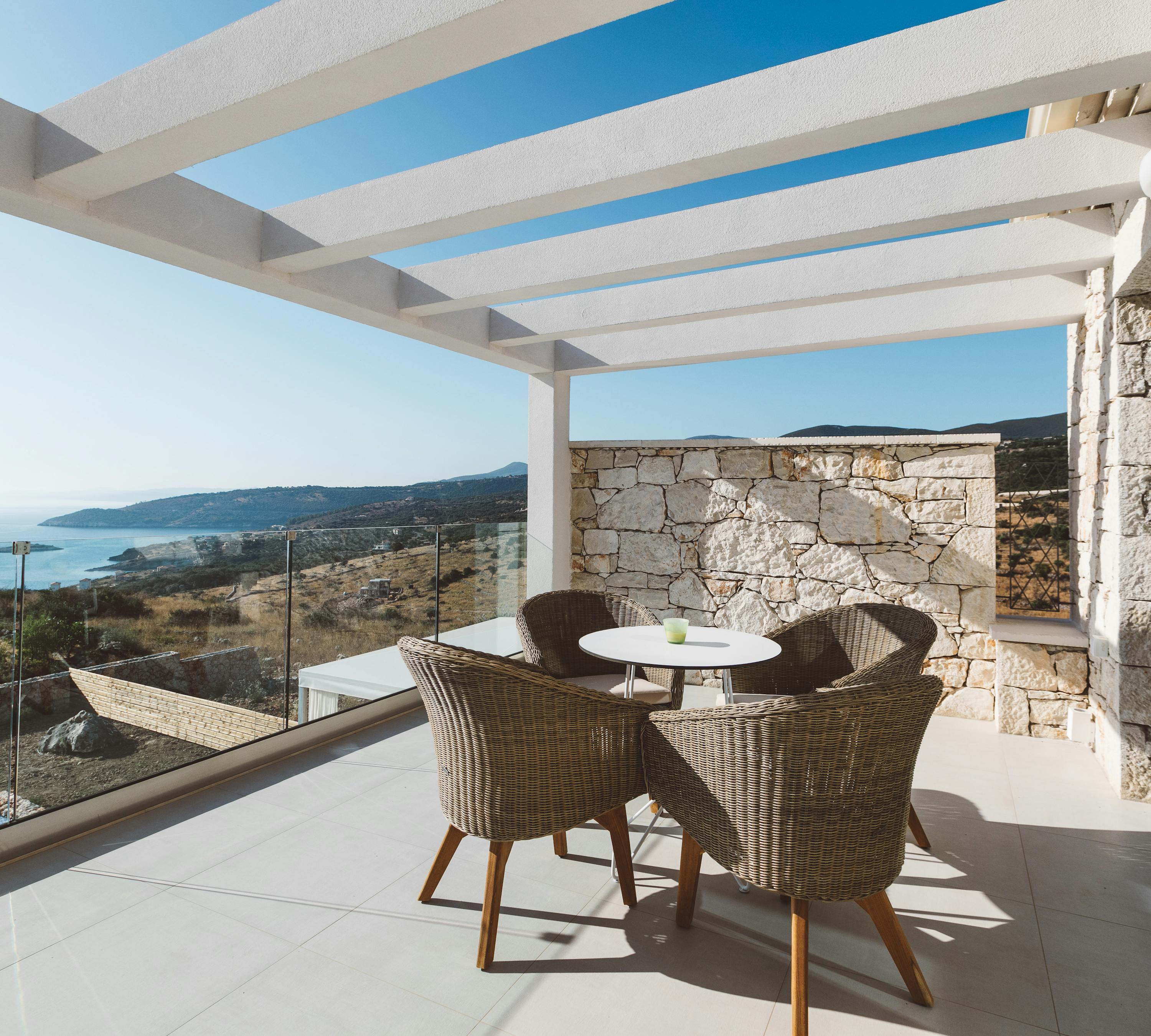 Covered terrace dining area with white pergola casting geometric shadows, wicker chairs around white table, stone accent wall, and glass-railed views of coastal hills.