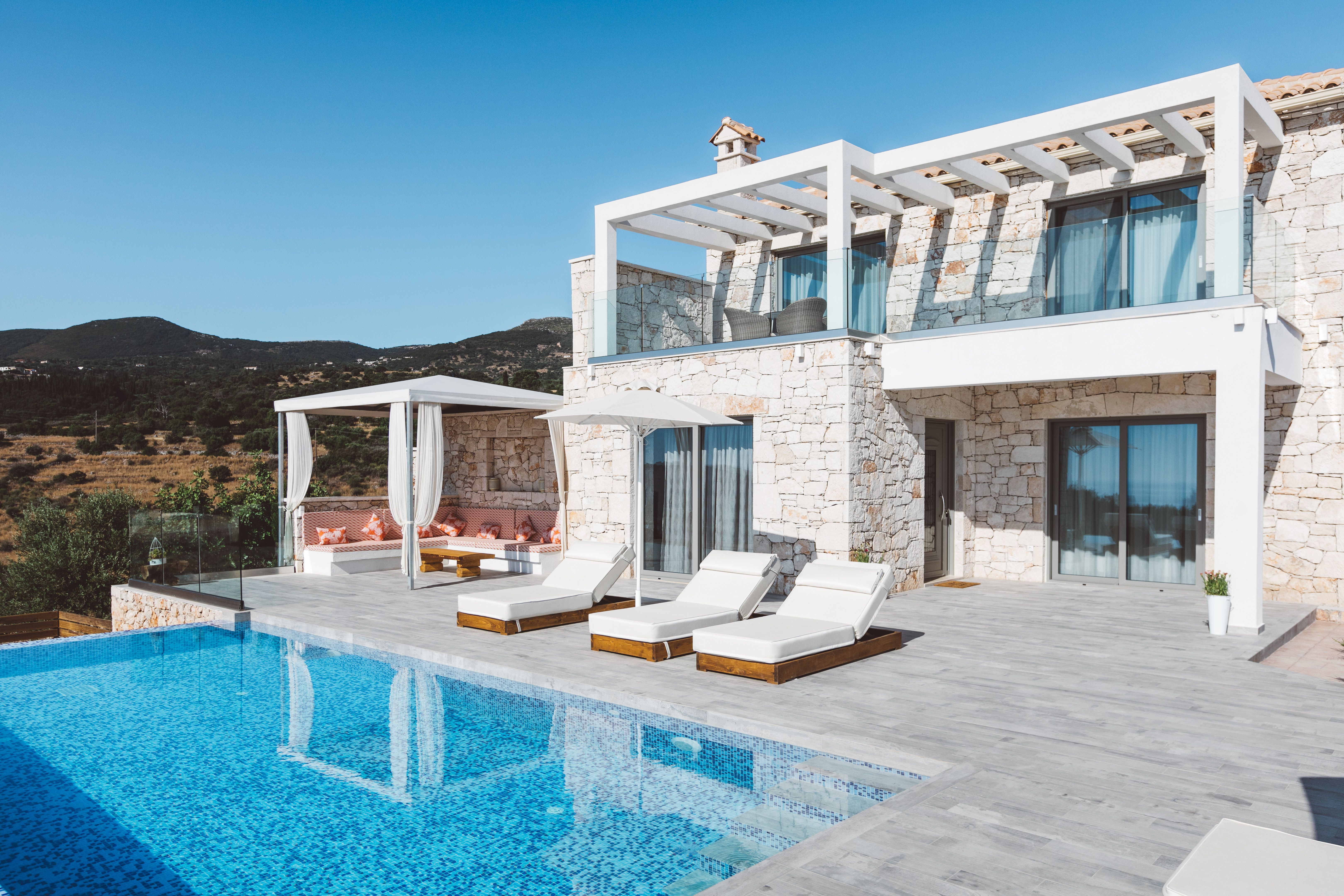 Two-story stone villa with white pergolas, infinity pool with submerged steps, white loungers, draped daybed pavilion, and mountain backdrop under clear blue sky.