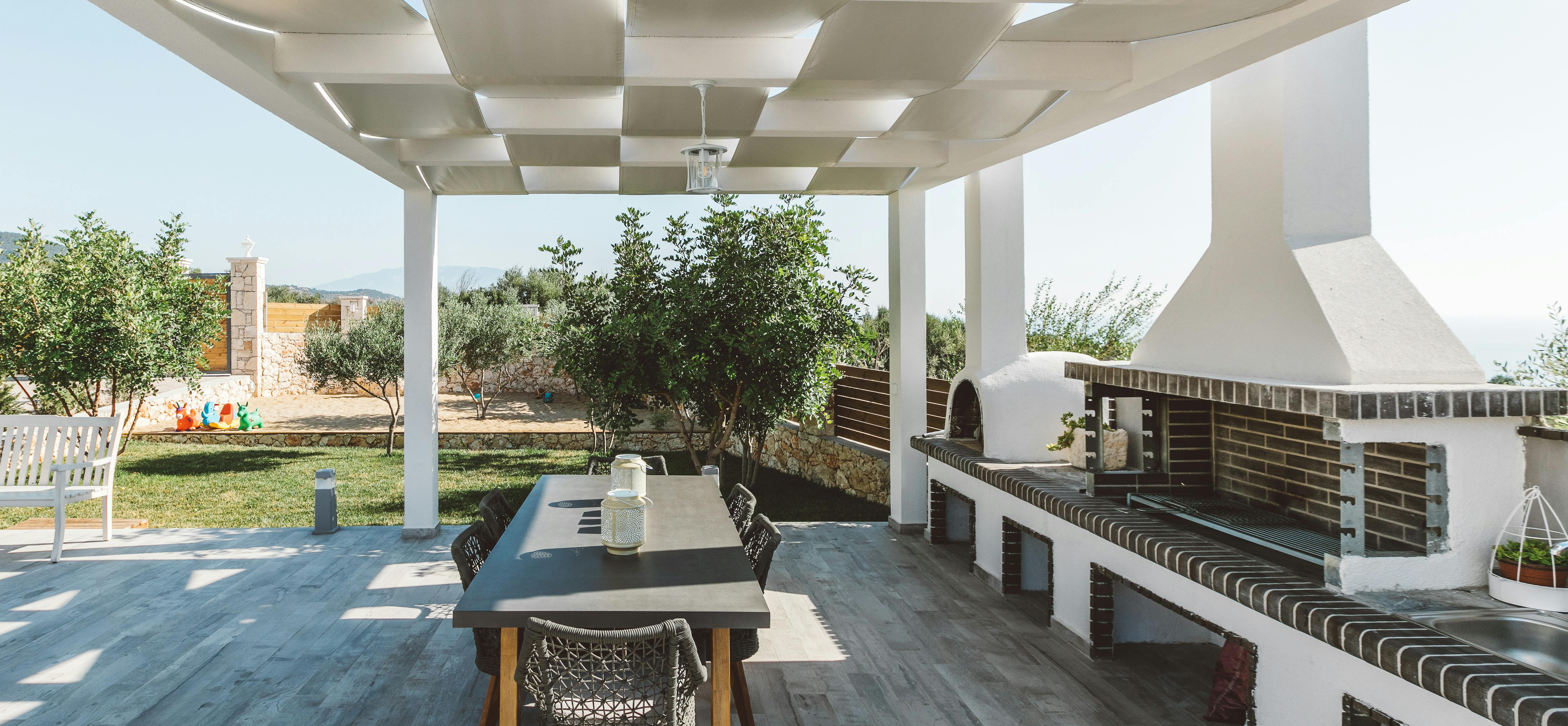 Covered outdoor dining area with white geometric pergola, dark wood table with woven chairs, white dome pizza oven, brick barbecue station, and views of lawn with olive trees.
