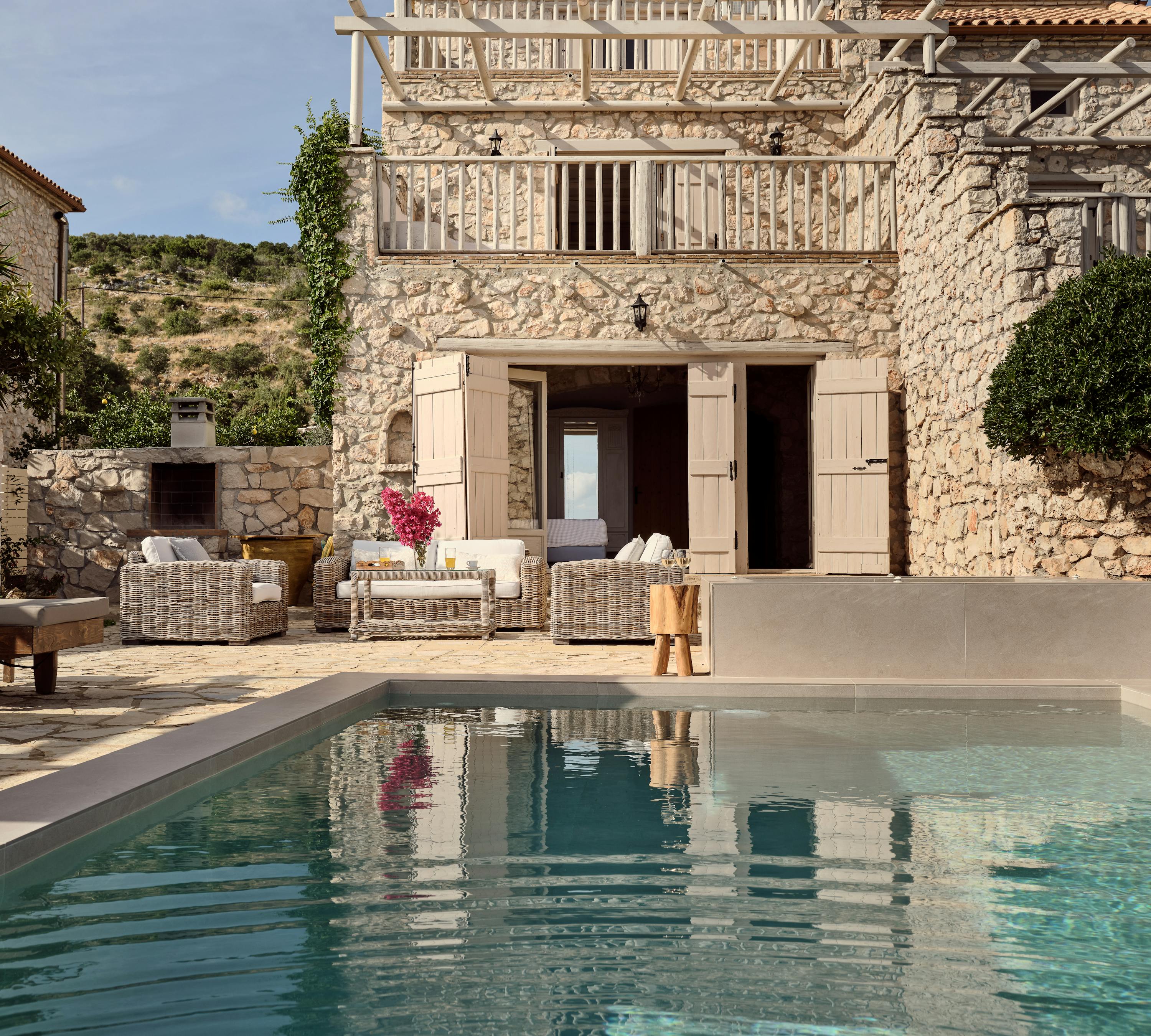 Two-story stone villa with upper balcony, pool terrace, and wicker outdoor furniture.