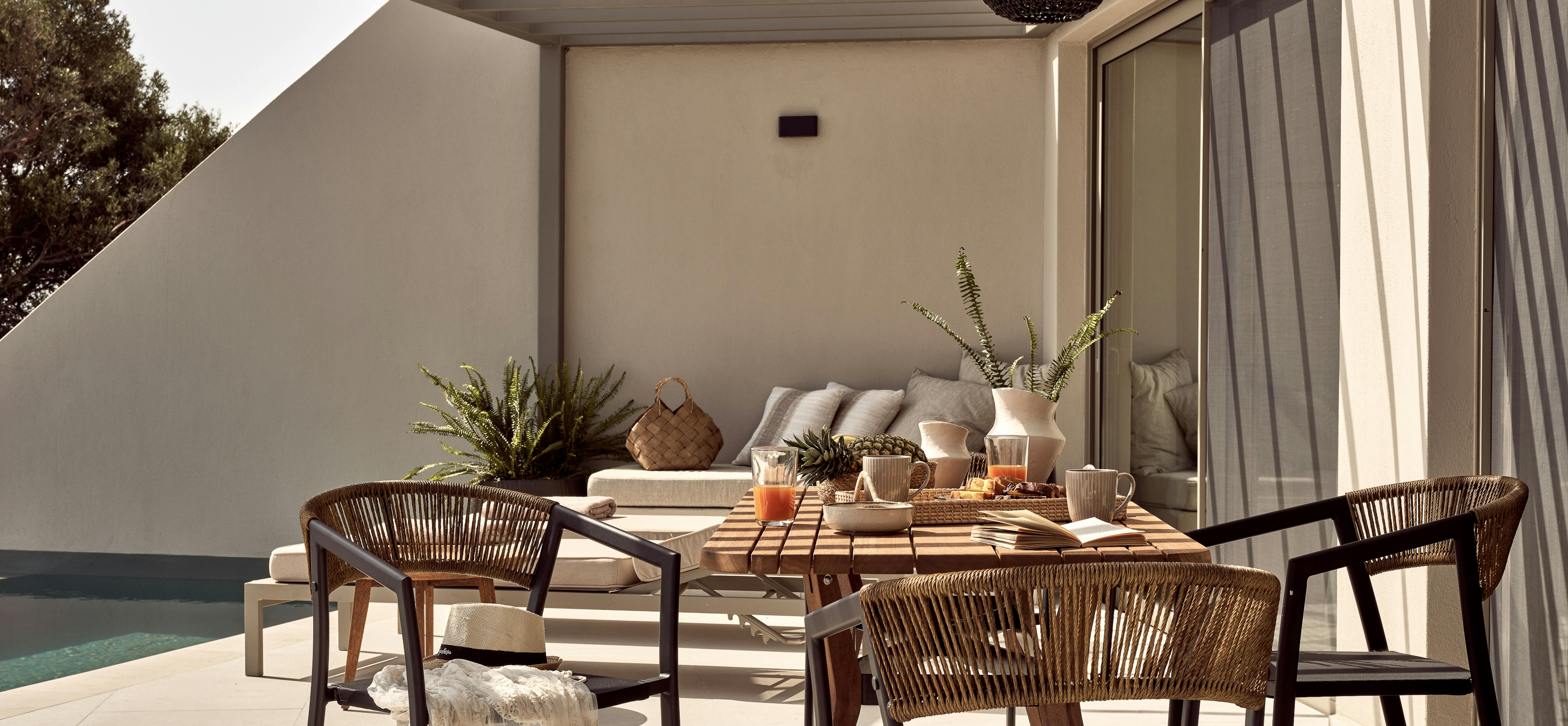 Covered terrace with breakfast setup on wooden table, woven chairs, and lounge seating by pool.