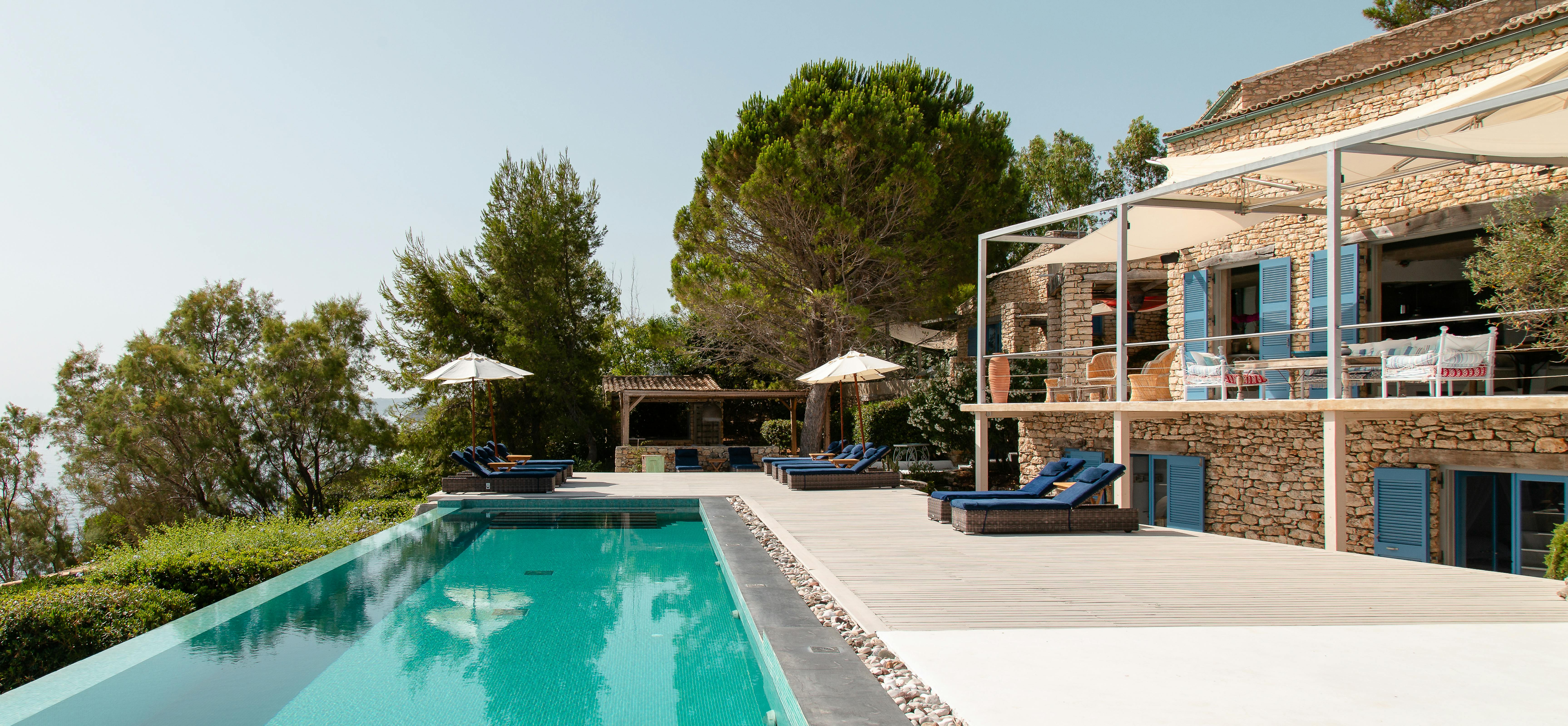 Mediterranean villa with infinity pool overlooking natural landscape. Stone architecture features blue shutters and modern outdoor furniture on terraced levels.
