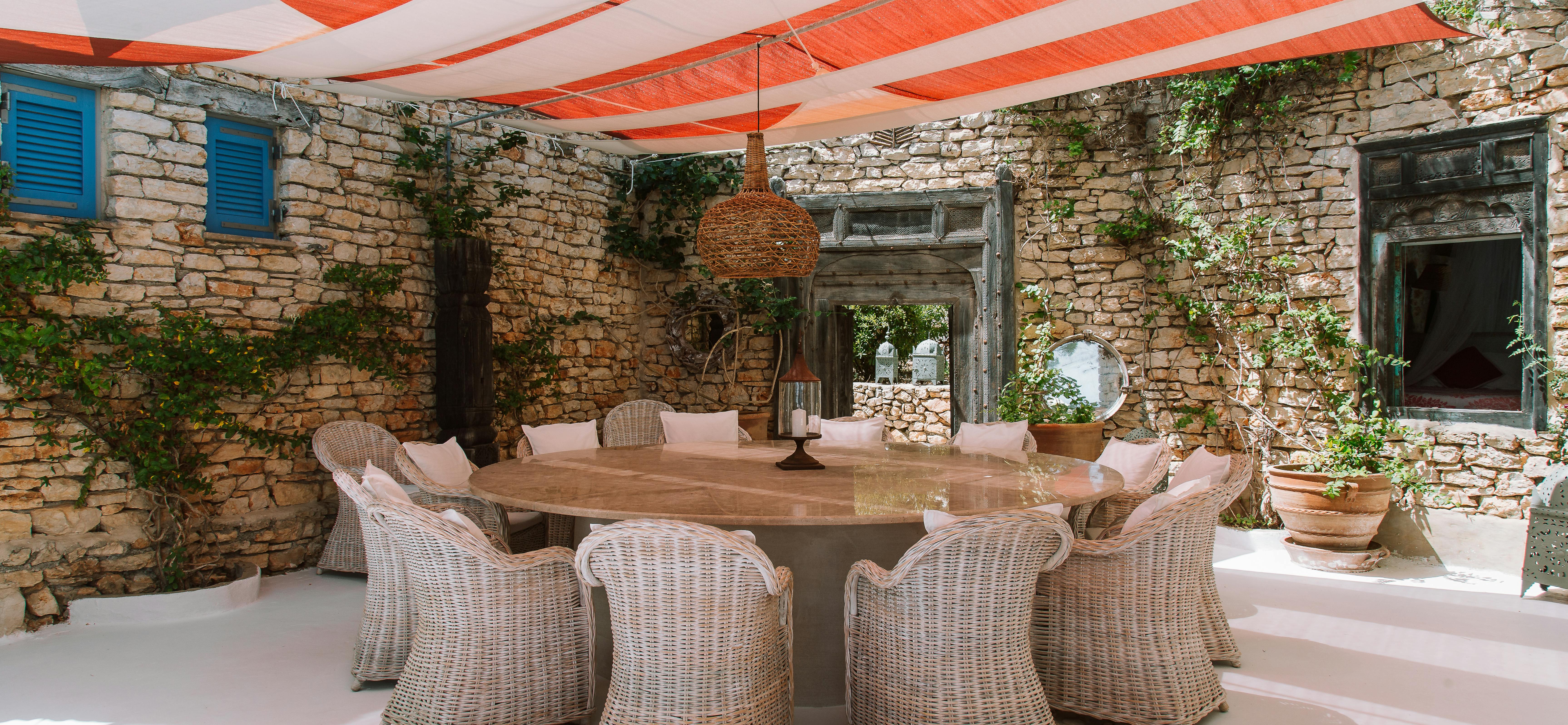 Covered outdoor dining area with wicker chairs around wooden table. Stone walls and orange fabric awning provide shade over the terrace space.