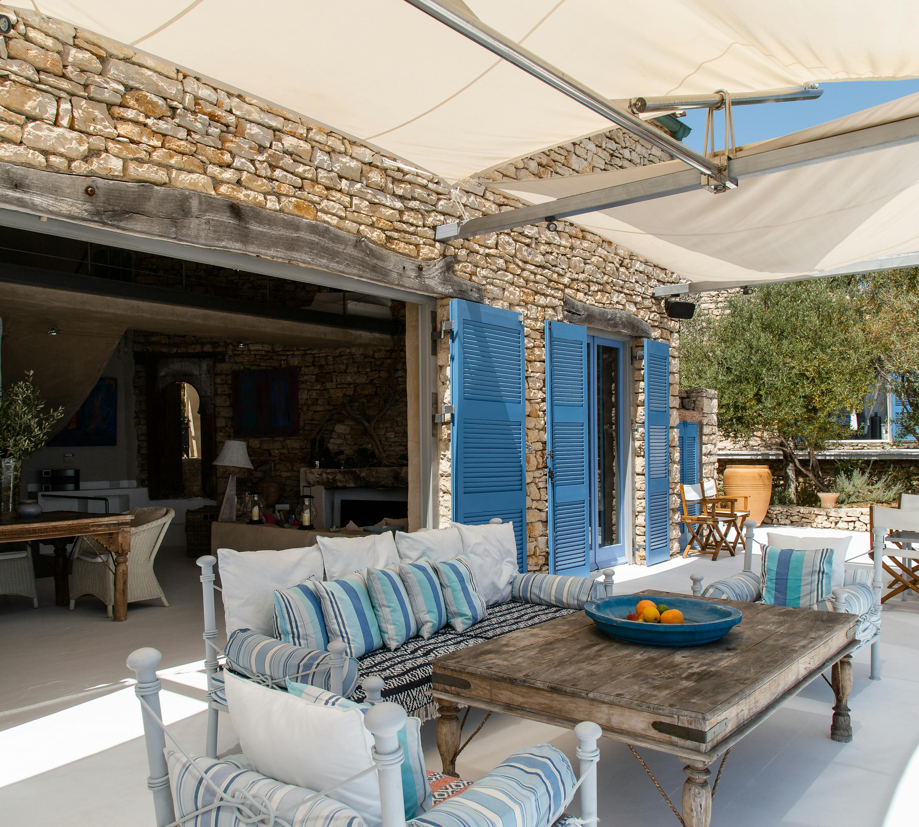 Covered outdoor living area with white furniture and blue striped cushions. Stone building with blue shutters forms backdrop to shaded terrace.