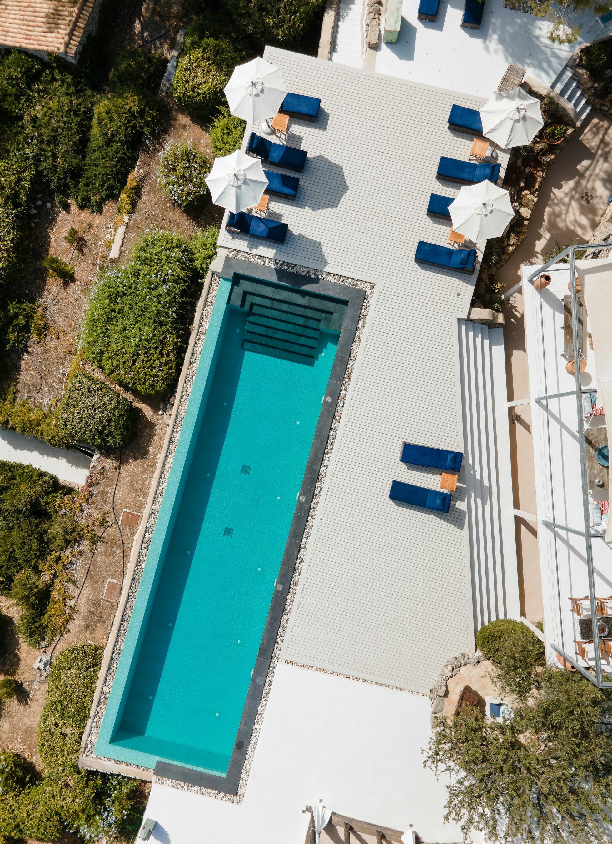 Aerial view of rectangular swimming pool with blue loungers arranged on white decking. Property surrounded by natural Mediterranean landscape and olive trees.
