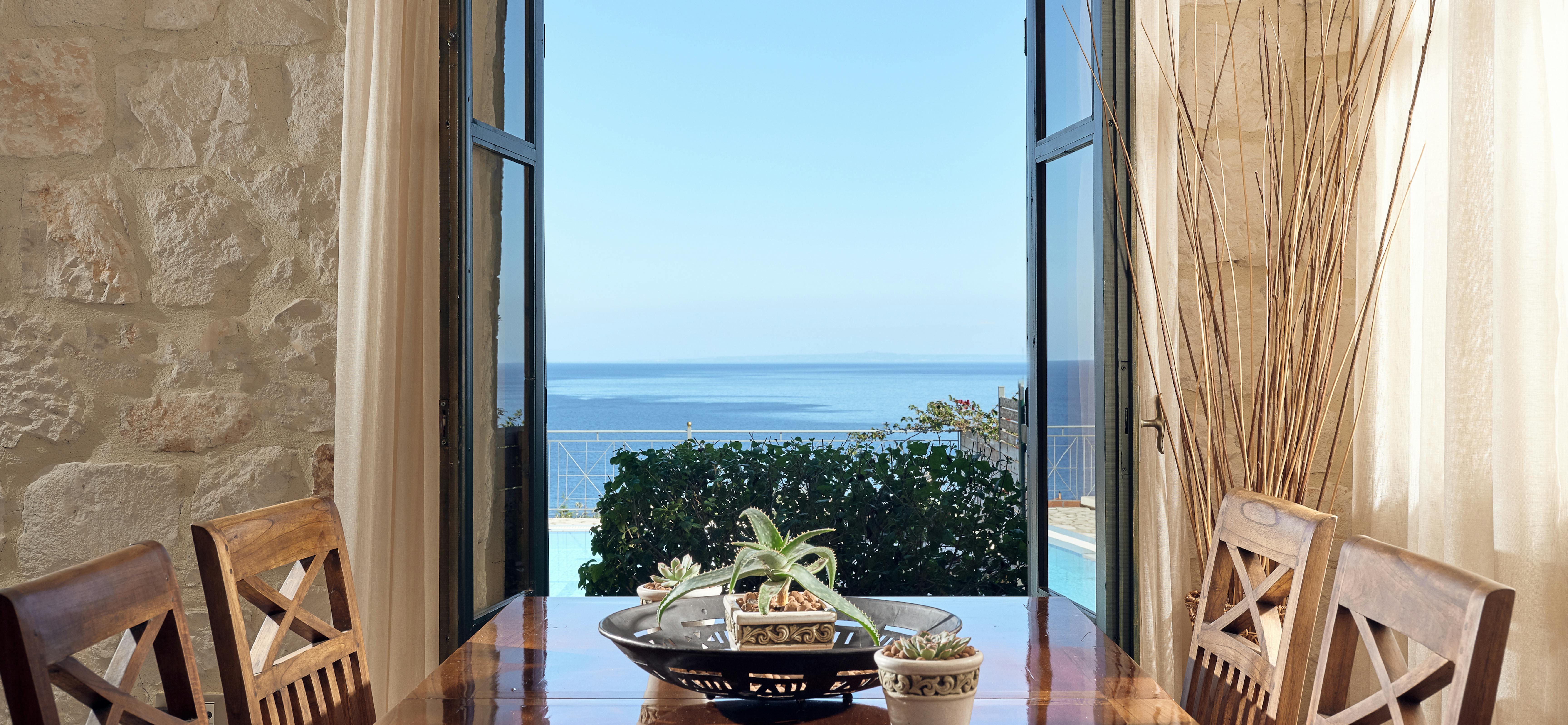Dining room with polished wood table and open French doors revealing pool and ocean vista.