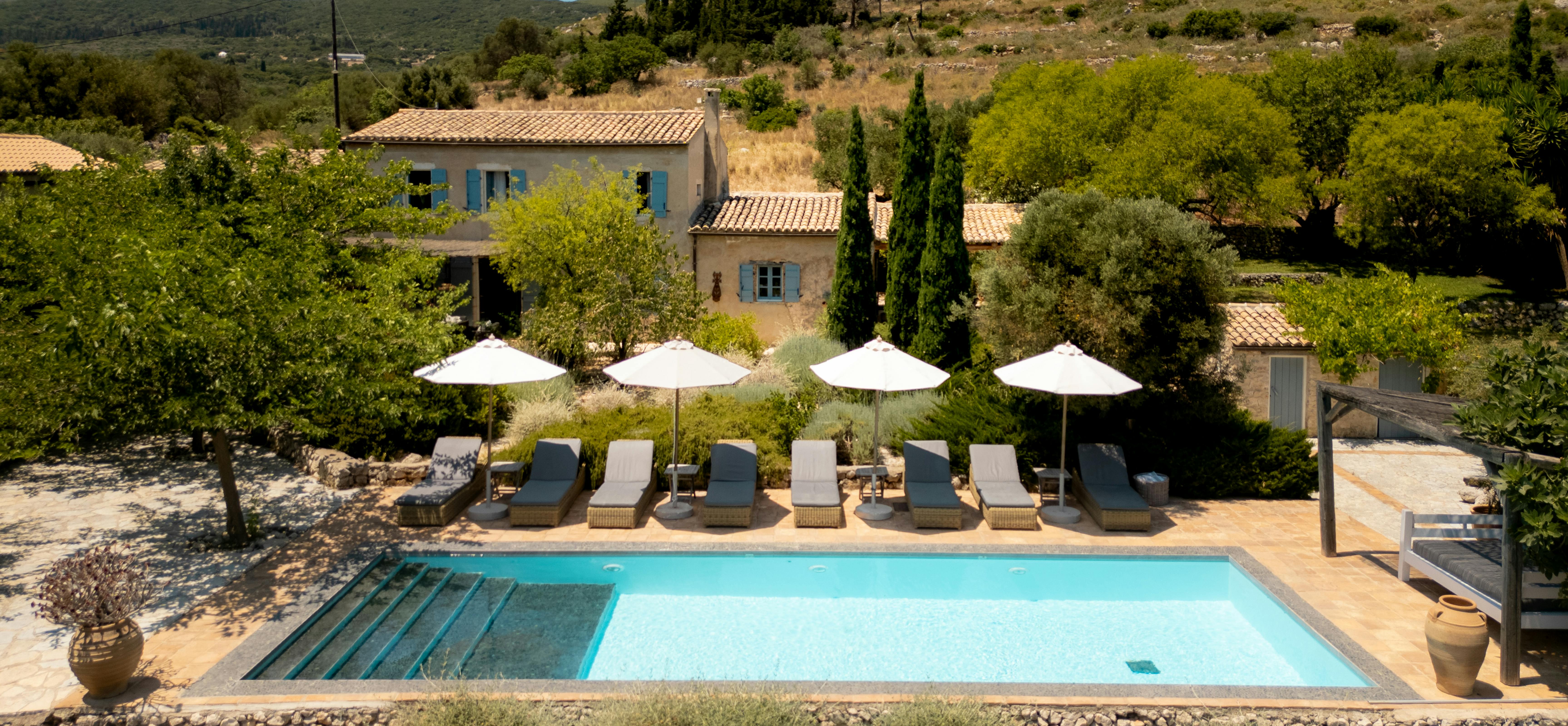 Elevated view of pool with beachside loungers and white umbrellas, overlooking terracotta-roofed villa amid olive groves and hills.