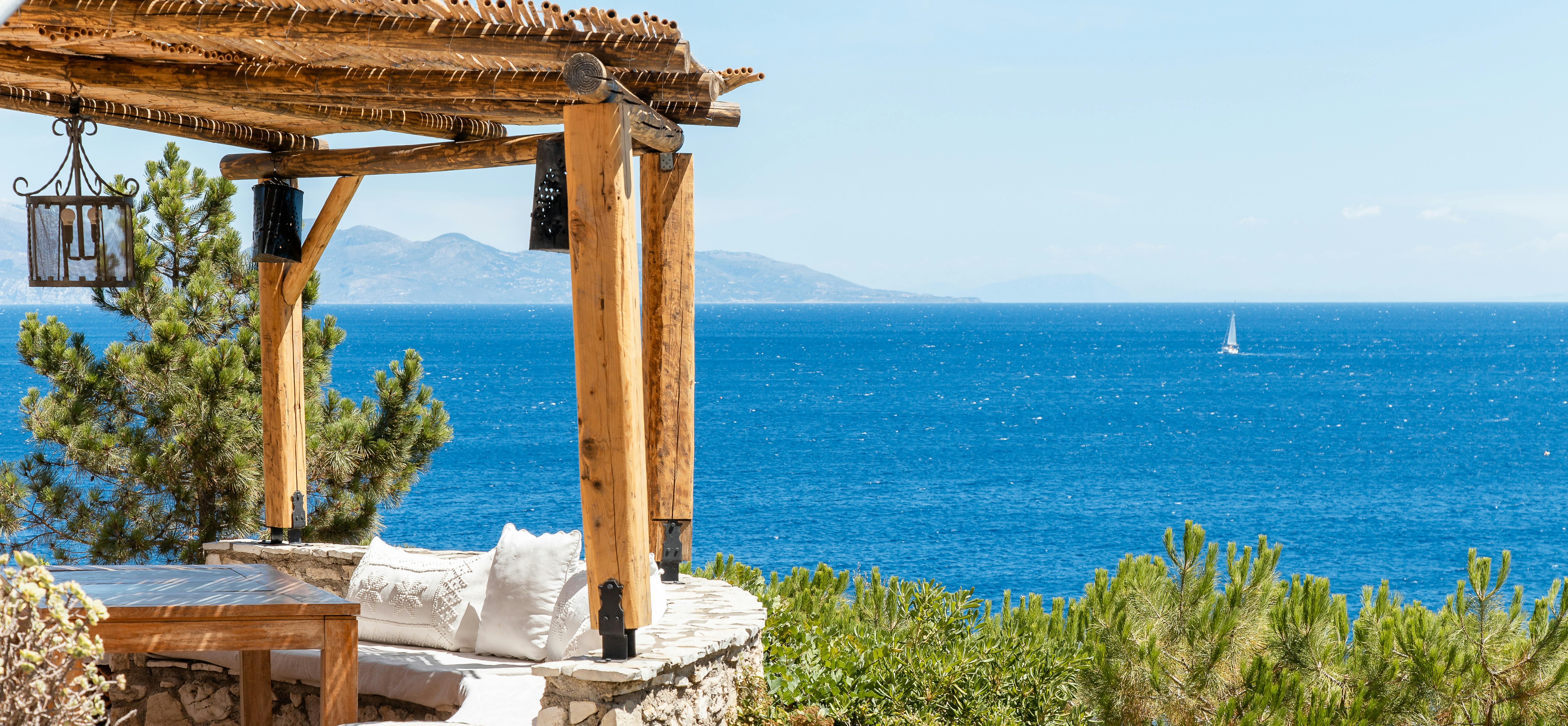 Rustic pergola with white daybed overlooking azure Mediterranean waters, with sailboat visible on horizon and distant mountain coastline.