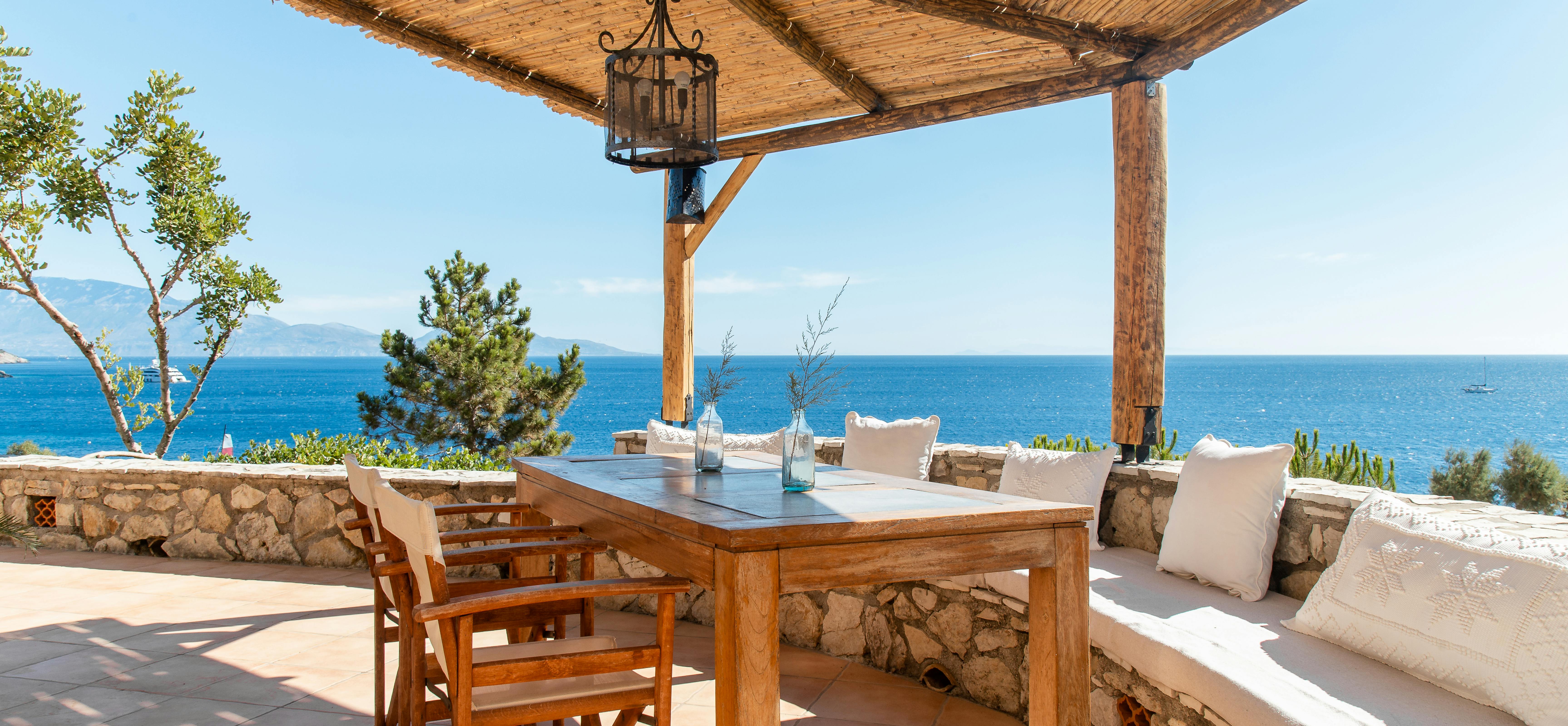 Covered terrace dining with reed ceiling, wooden table with built-in stone seating, and panoramic sea views framed by lantern.