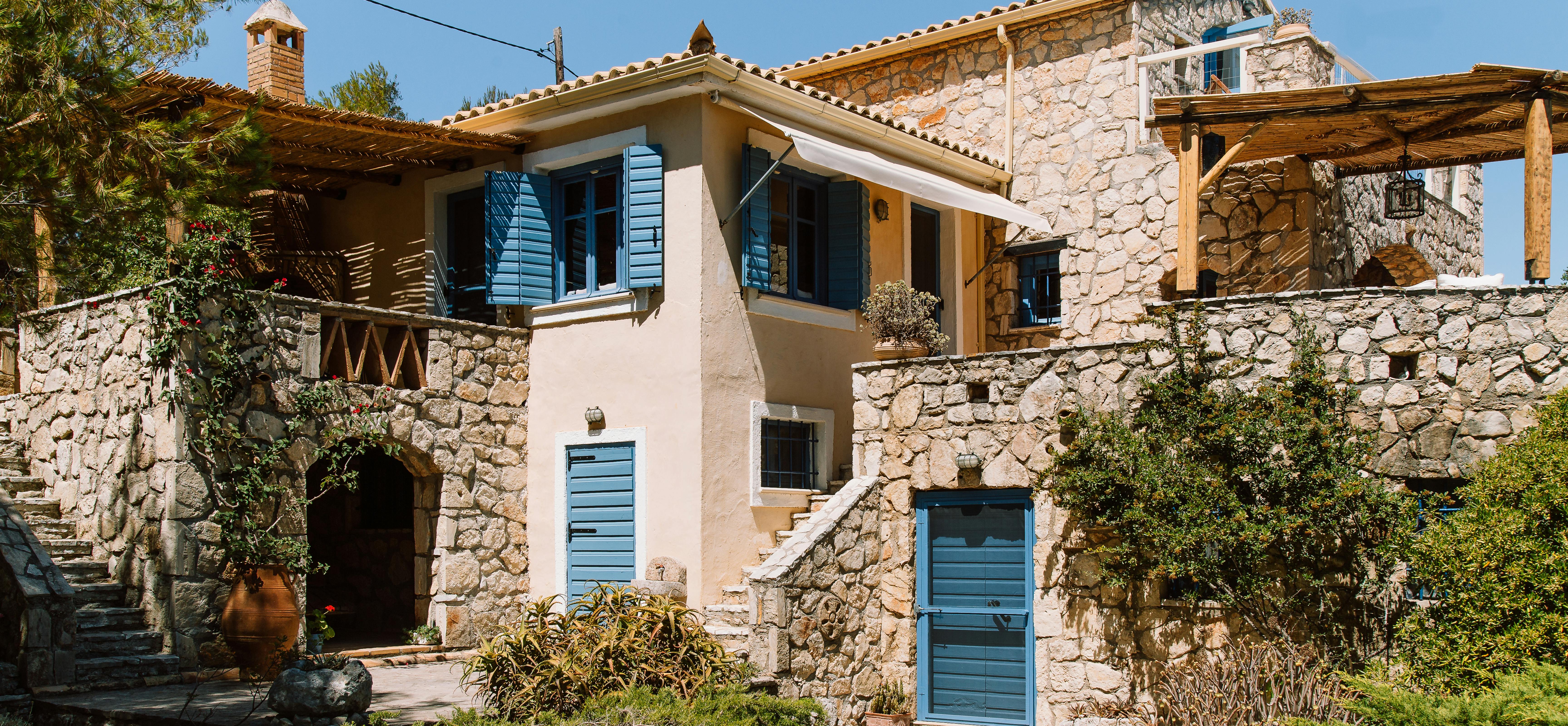 Two-story stone cottage with blue shutters, terracotta roofs, wooden pergolas, and terraced garden with terracotta planters.