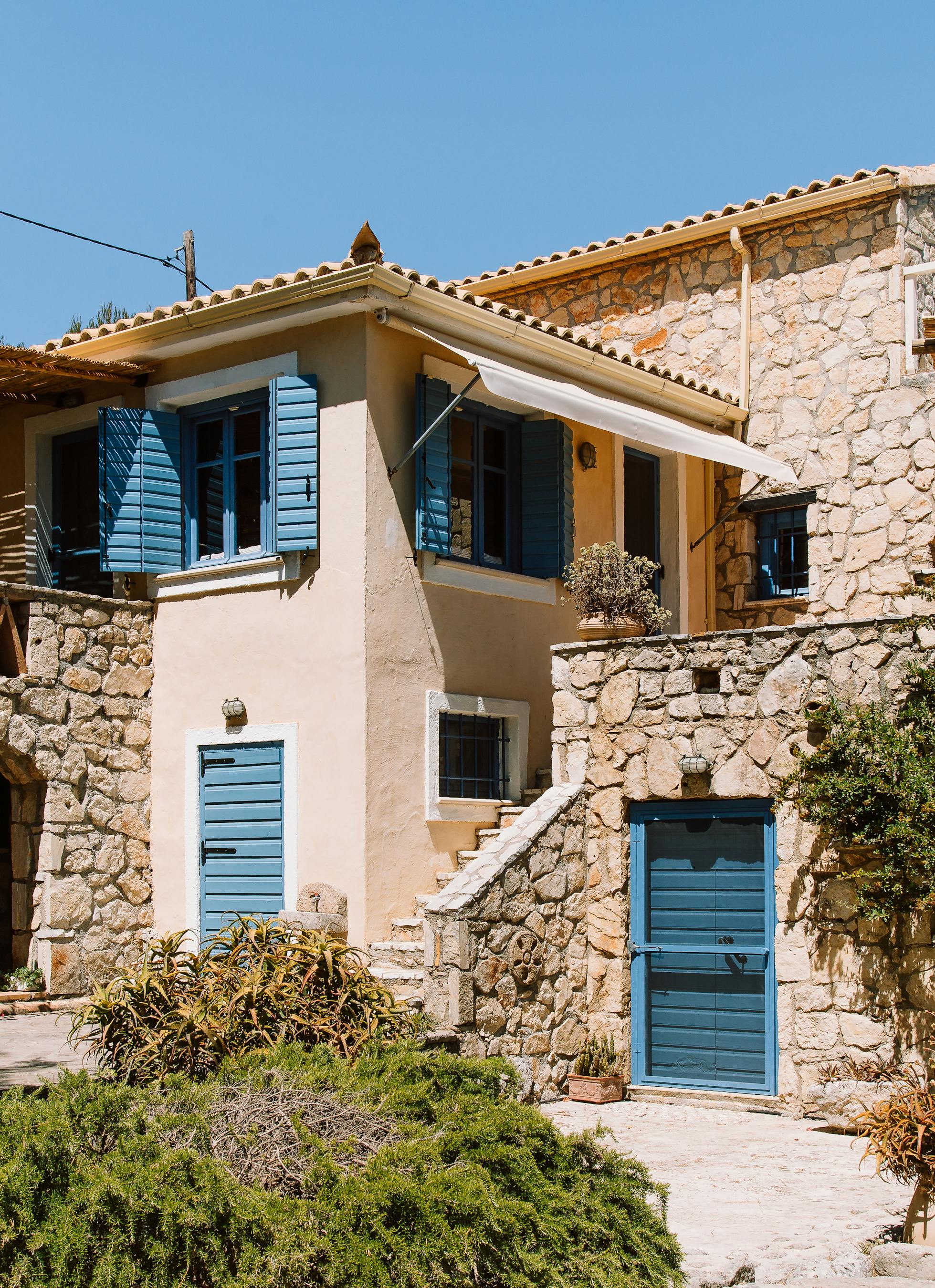 Two-story stone cottage with blue shutters, terracotta roofs, wooden pergolas, and terraced garden with terracotta planters.