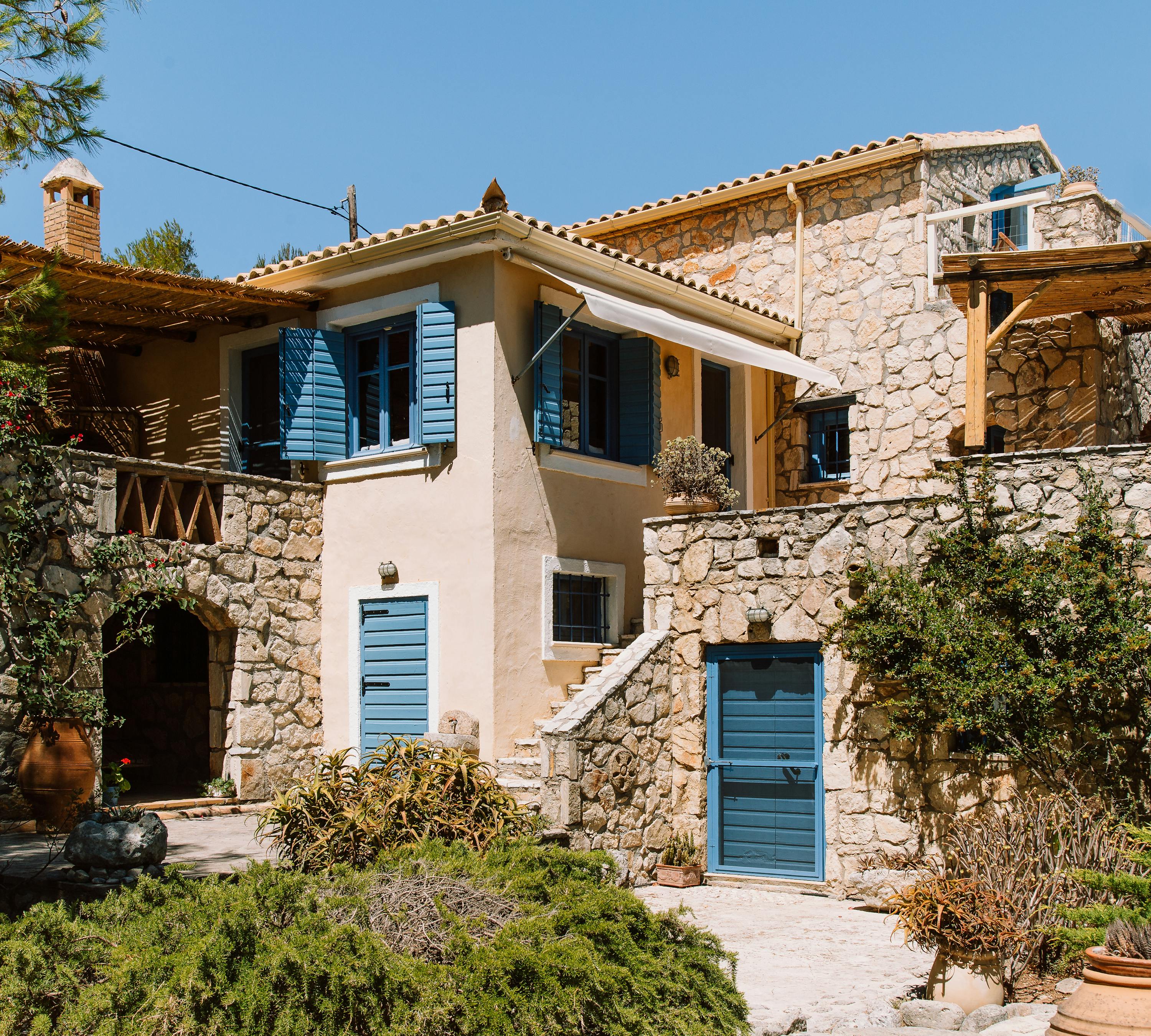 Two-story stone cottage with blue shutters, terracotta roofs, wooden pergolas, and terraced garden with terracotta planters.