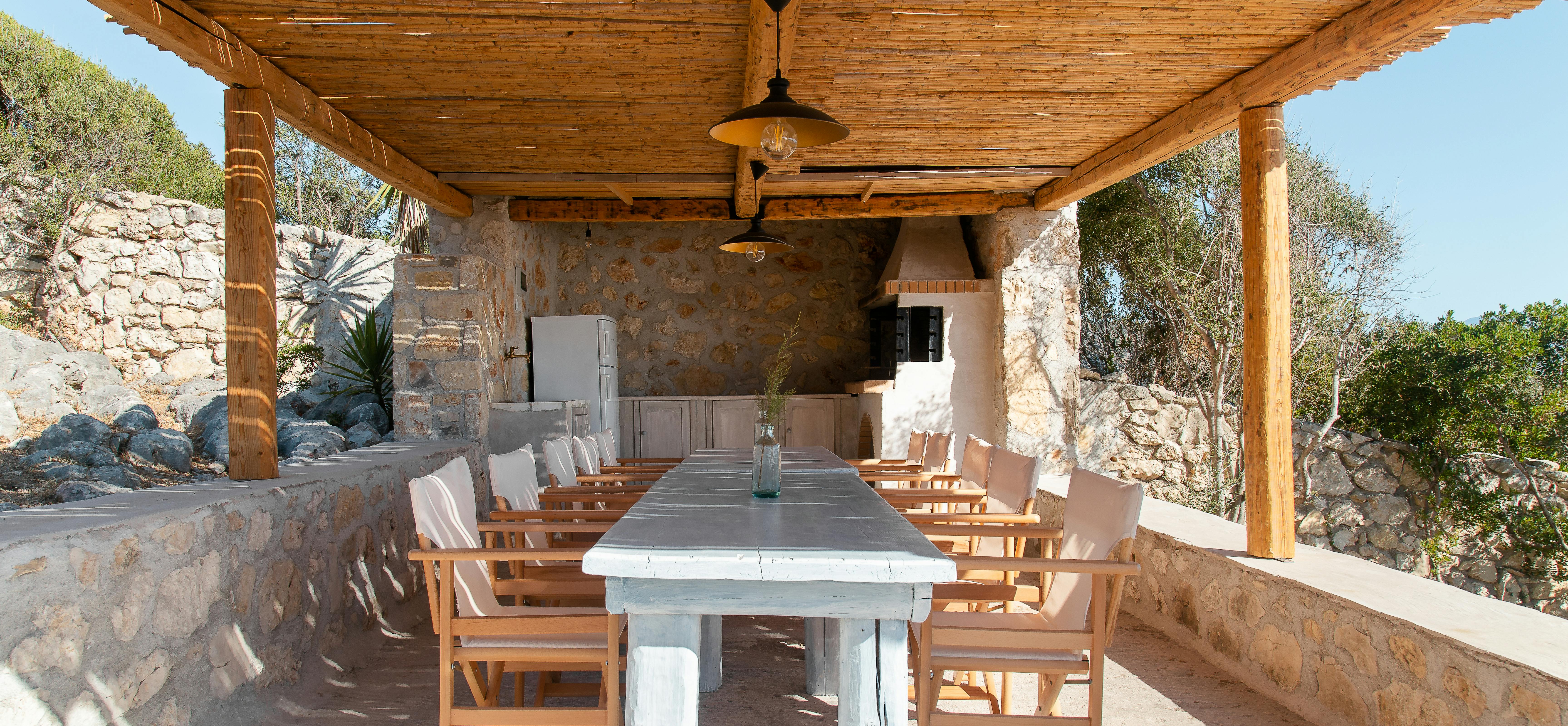 Open-air dining pavilion with stone walls, whitewashed table, wooden chairs, and built-in grill beneath reed roof canopy.