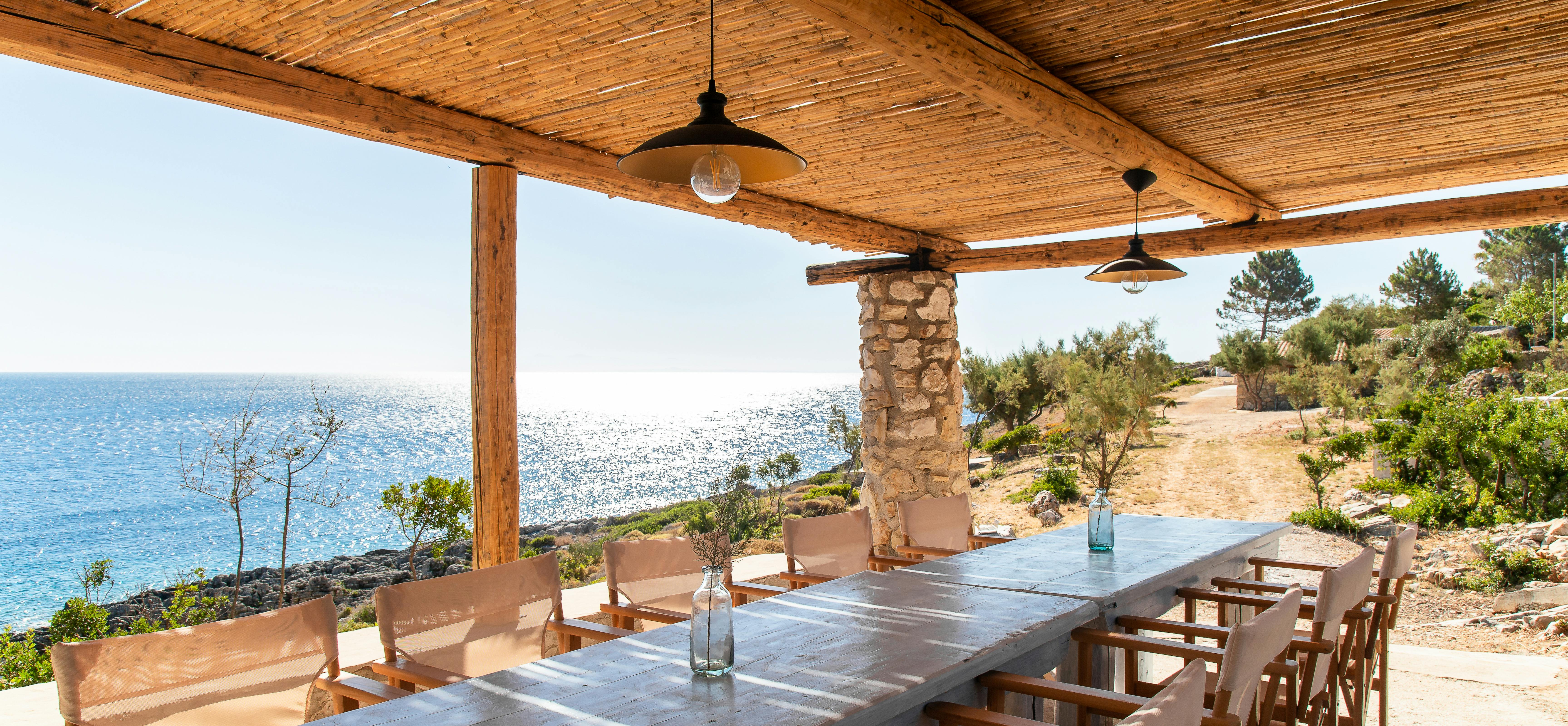 Seaside dining terrace with reed ceiling, stone column, long wooden tables, and unobstructed Mediterranean coastal panorama.