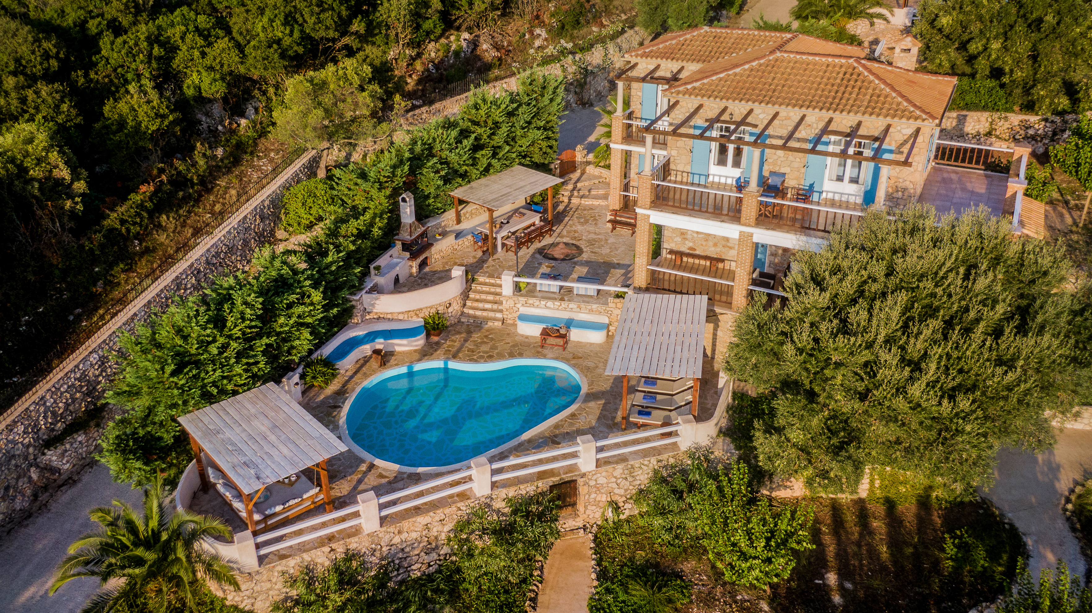 Aerial perspective of hillside villa with curved pool, multiple wooden pergolas, terraced levels, and olive grove surroundings.