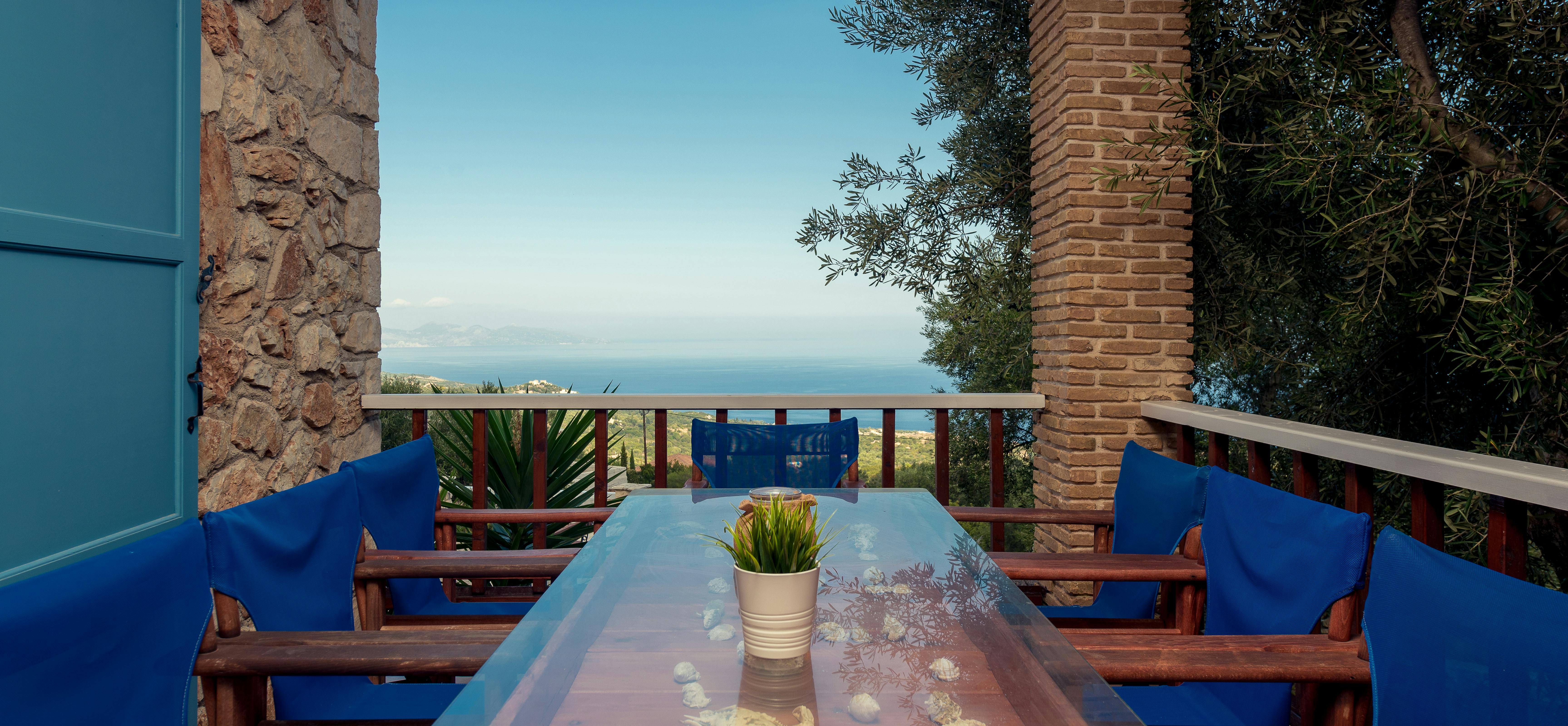 Covered terrace dining with blue cushioned chairs, glass table, brick columns, and panoramic coastal mountain views.
