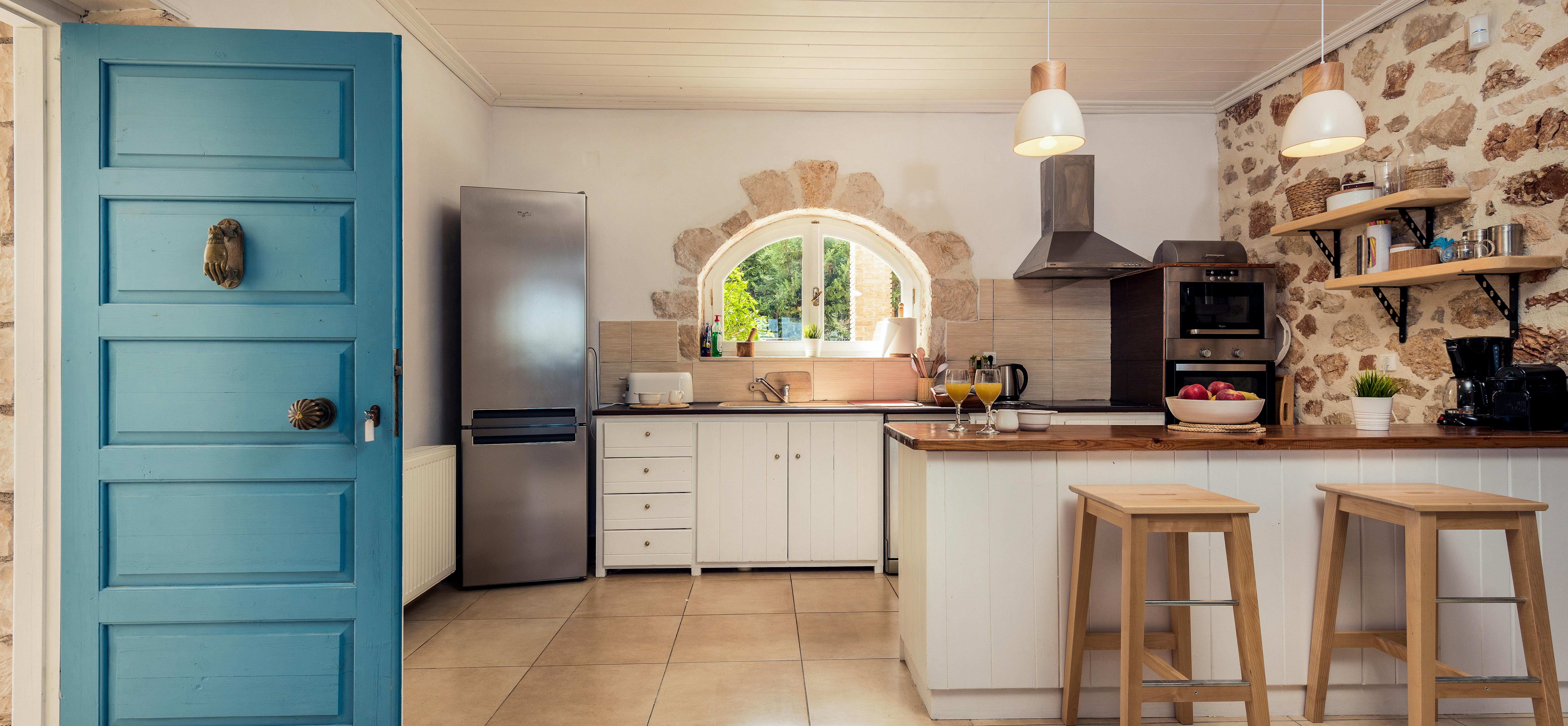 Bright kitchen with exposed stone wall, white cabinets, wooden counters, blue door, and arched window overlooking garden.
