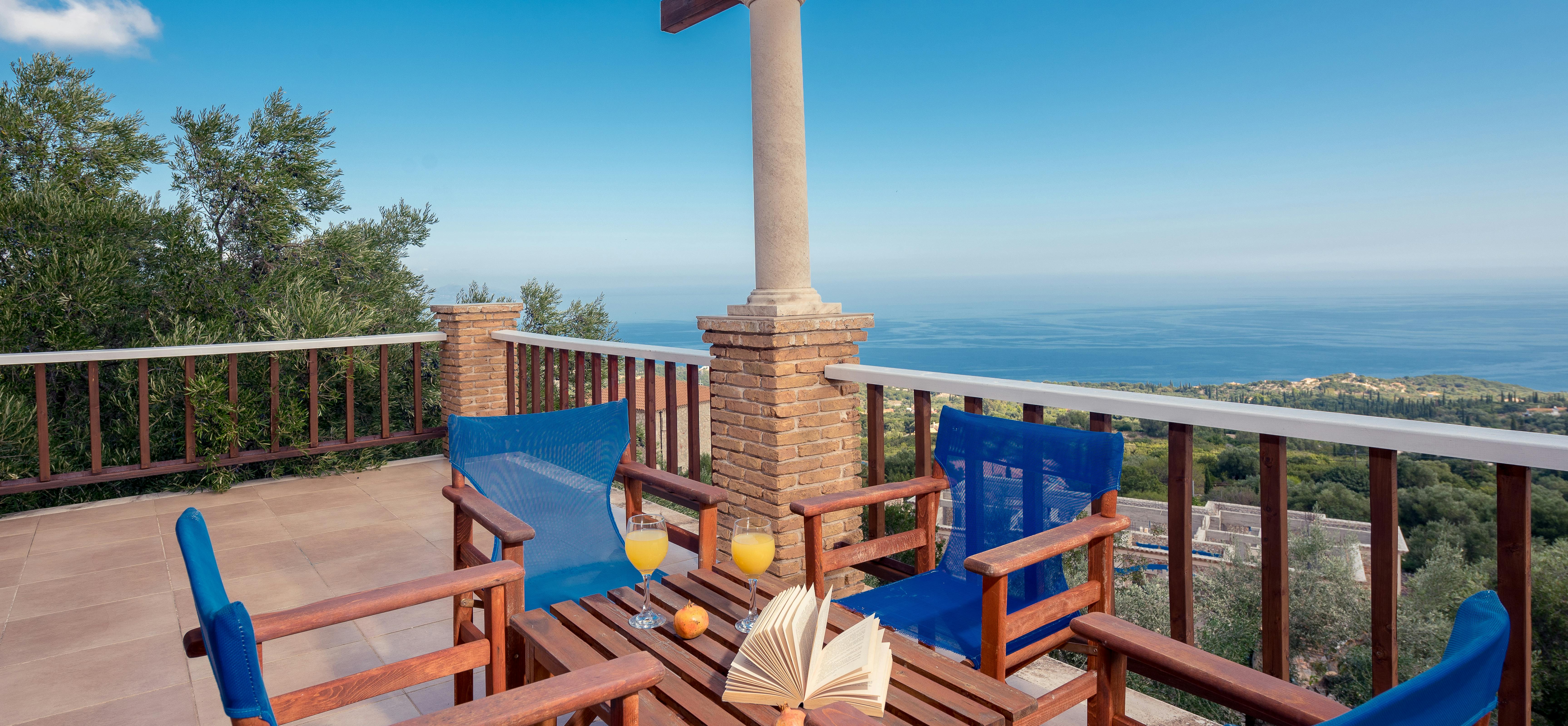 A covered terrace with blue-cushioned wooden chairs frames a spectacular sunset view over the sea and coastal landscape.