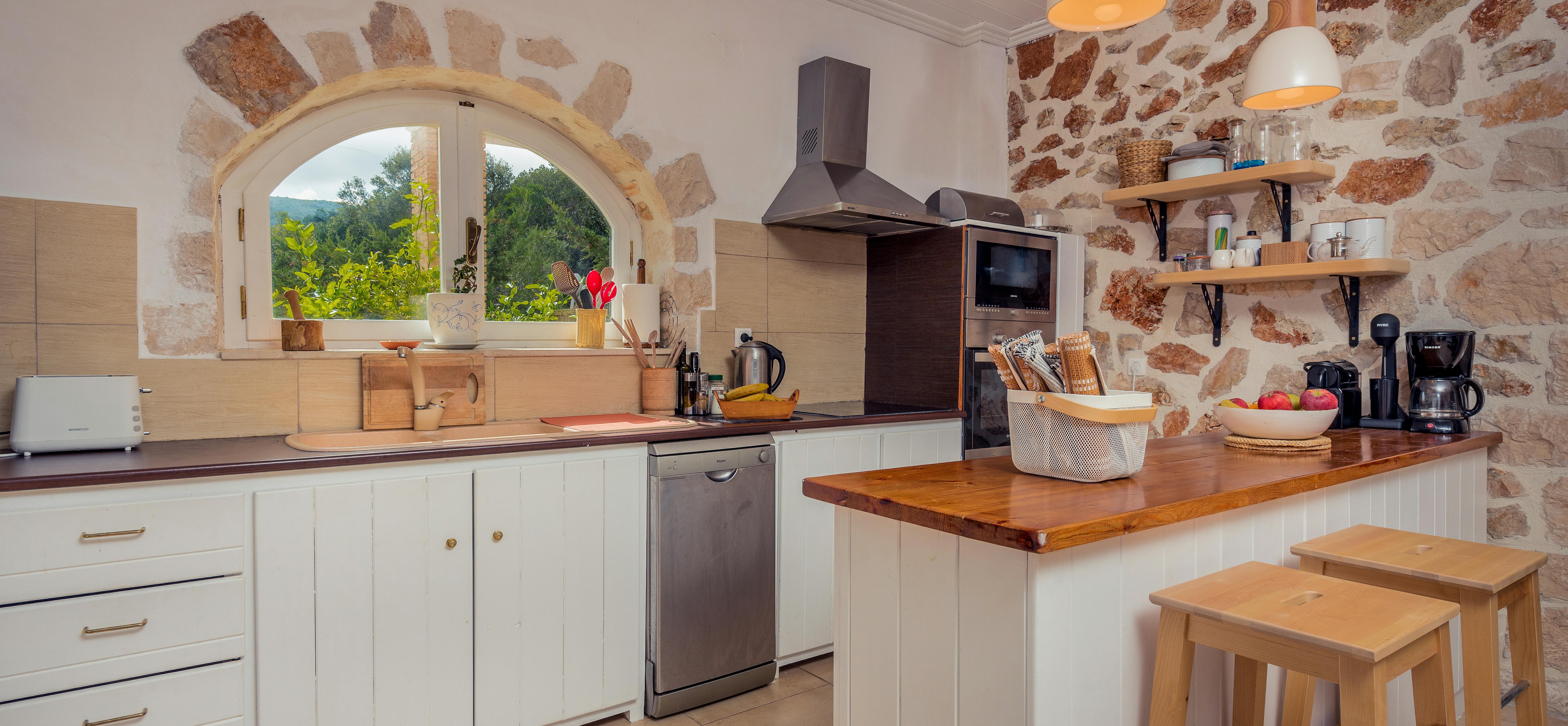A rustic Mediterranean kitchen features exposed stone walls, an arched window with garden views, white cabinets with wooden countertops, and a breakfast bar with two wooden stools.