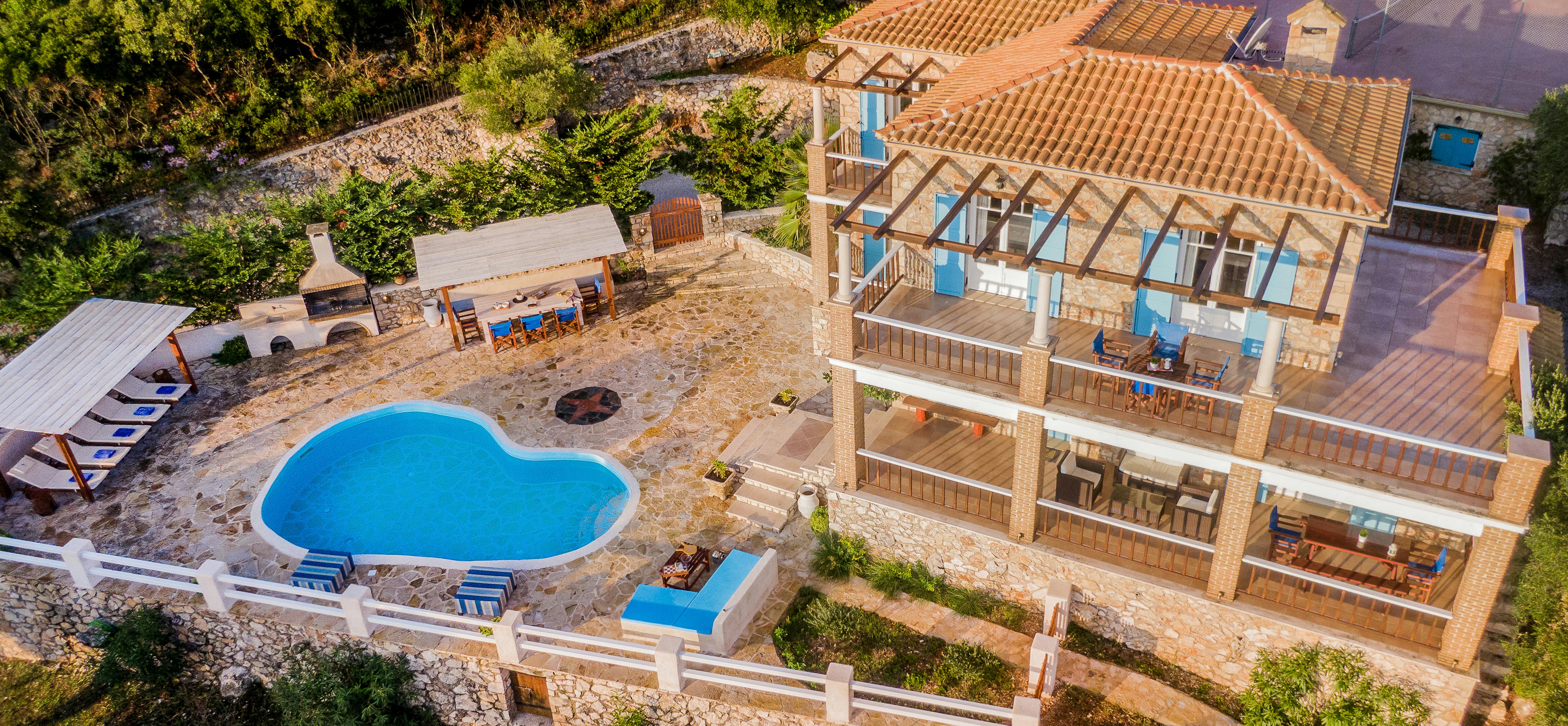 Aerial view of a two-story stone villa with terracotta roof and wooden pergola structures, featuring multiple balconies with blue accents, a kidney-shaped pool, and an outdoor dining area on a terraced hillside property.