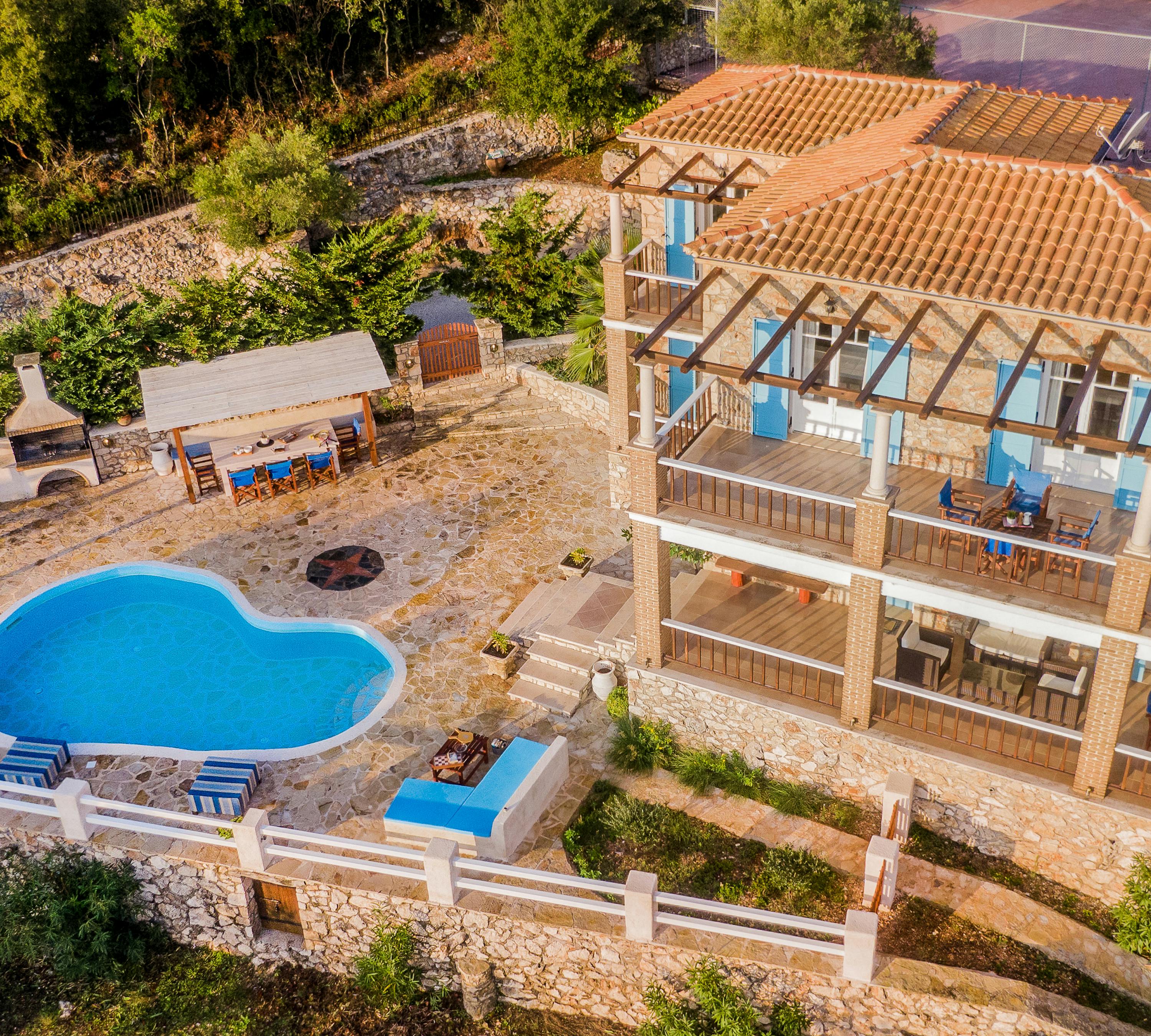 Aerial view of a two-story stone villa with terracotta roof and wooden pergola structures, featuring multiple balconies with blue accents, a kidney-shaped pool, and an outdoor dining area on a terraced hillside property.