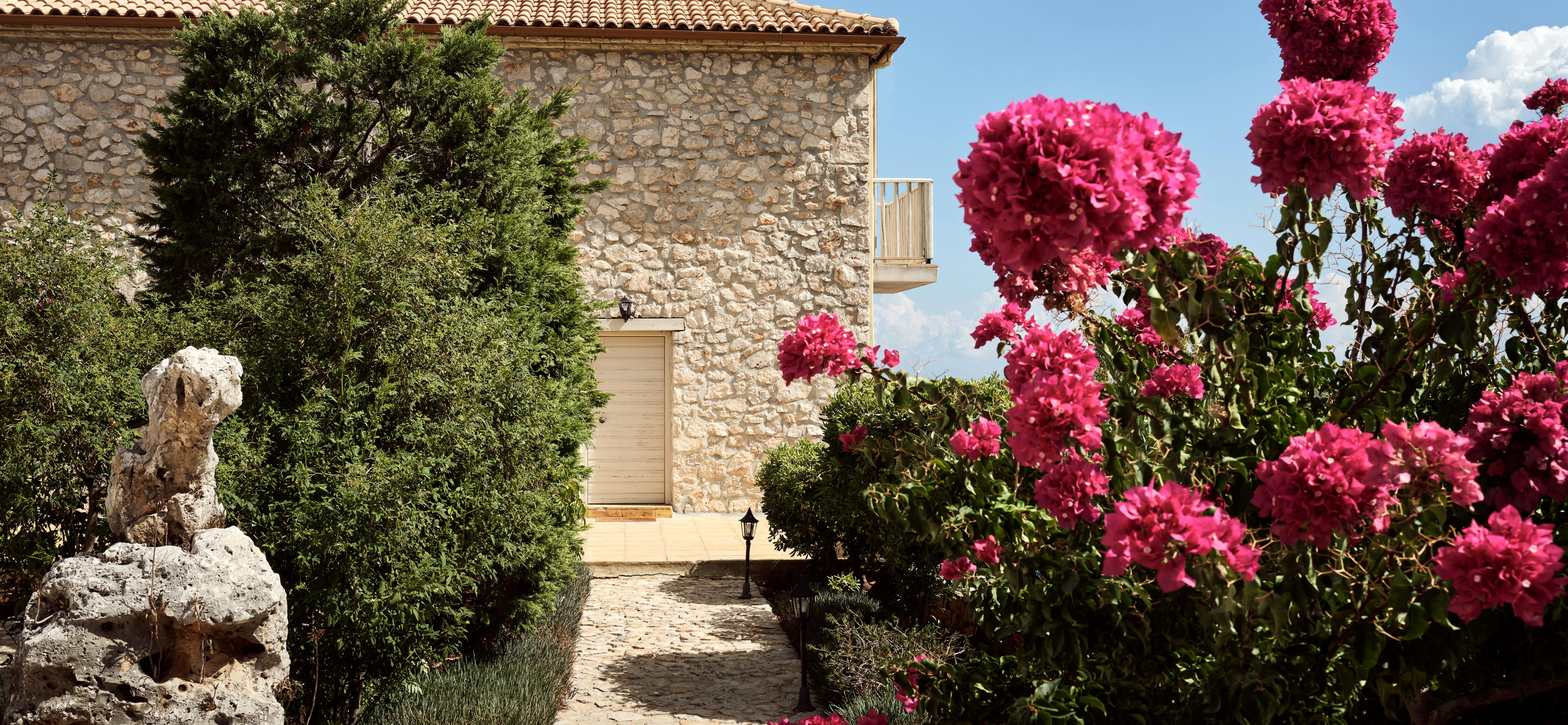 Stone Mediterranean villa entrance with vibrant pink bougainvillea blooms framing a cobblestone pathway leading to a cream-coloured door under terracotta roof tiles.